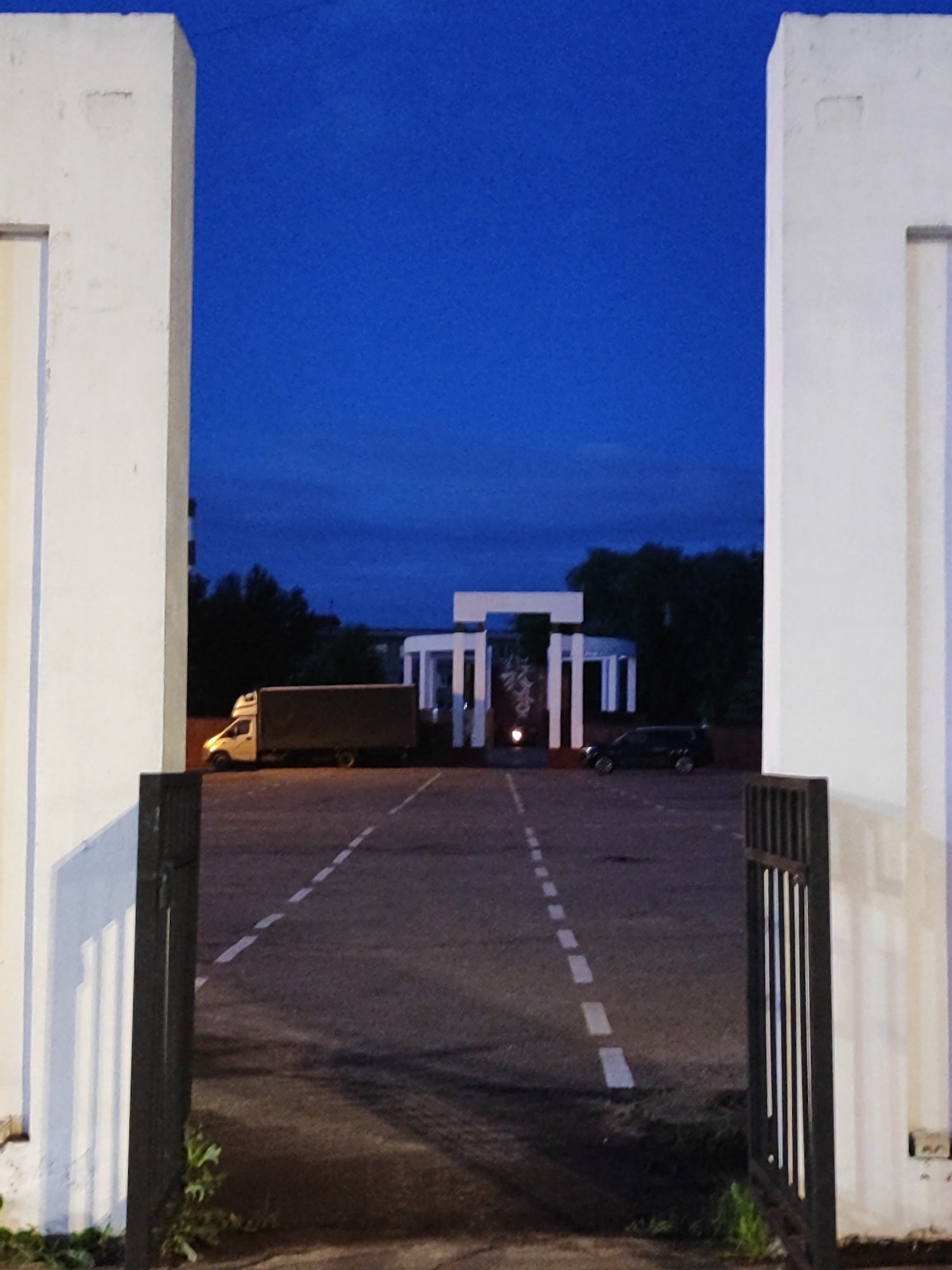 At dusk, an outdoor scene is captured through a wide archway framing a dark blue sky. Two large, white, rectangular pillars stand on either side of the archway. In the middle ground, a white, modern, open-air structure with multiple pillars and horizontal beams is visible, illuminated by a single light source in its center. To the left of this structure, a white box truck with a gray cargo area is parked on the asphalt. To the right, a dark-colored SUV is also parked. The ground is paved asphalt with dashed white lines indicating a roadway or parking area. A black metal gate with vertical bars is partially open on the right side of the foreground. Trees form a dark silhouette in the background behind the central structure.