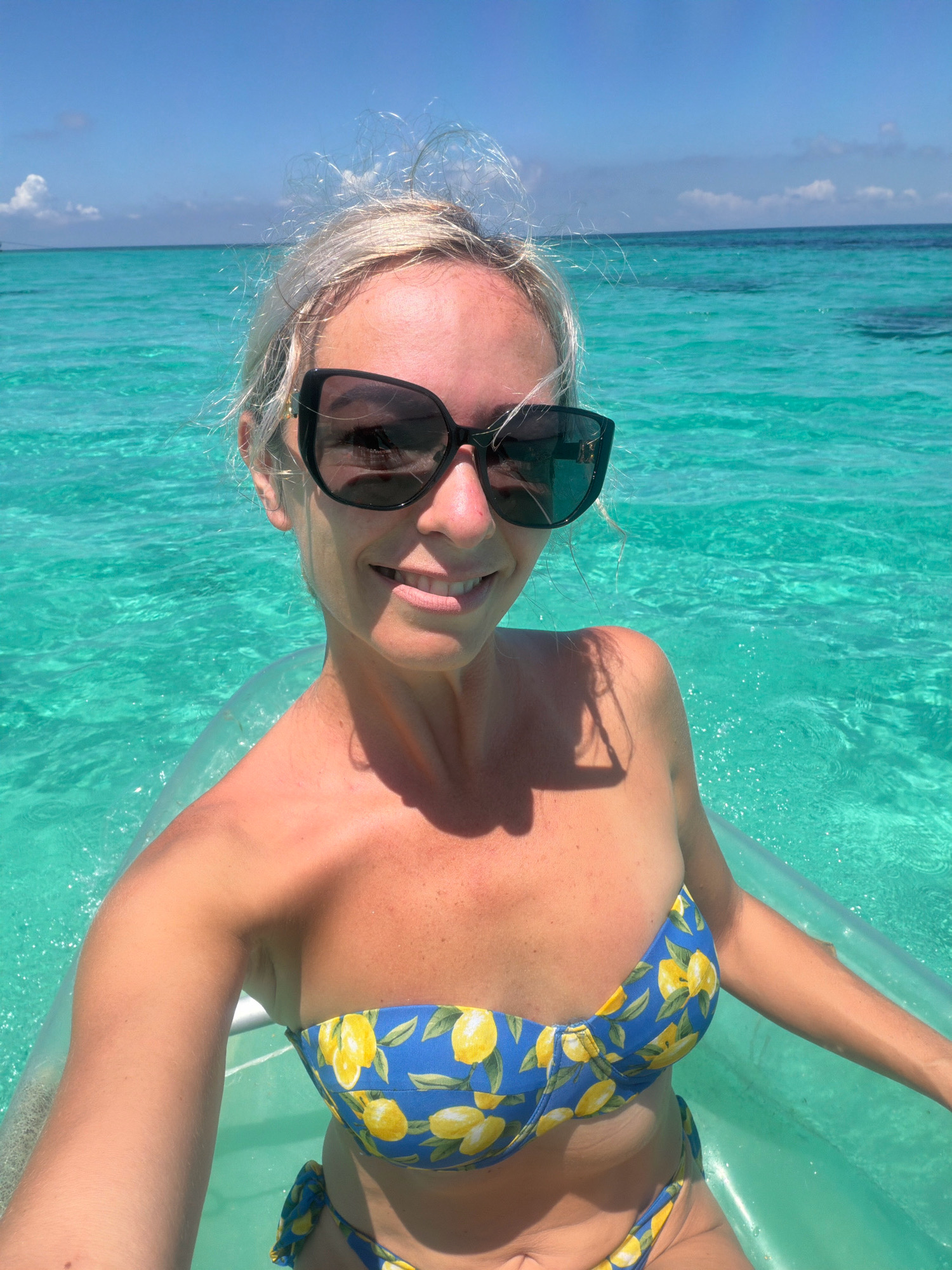A woman with blonde hair and sunglasses is taking a selfie in the crystal clear waters of Boracay, Malay, Philippines. She is wearing a blue bikini with a lemon print and is sitting in a clear float. The water is turquoise and calm, with the sky a clear blue above. The sun is shining brightly, casting shadows on the water.
