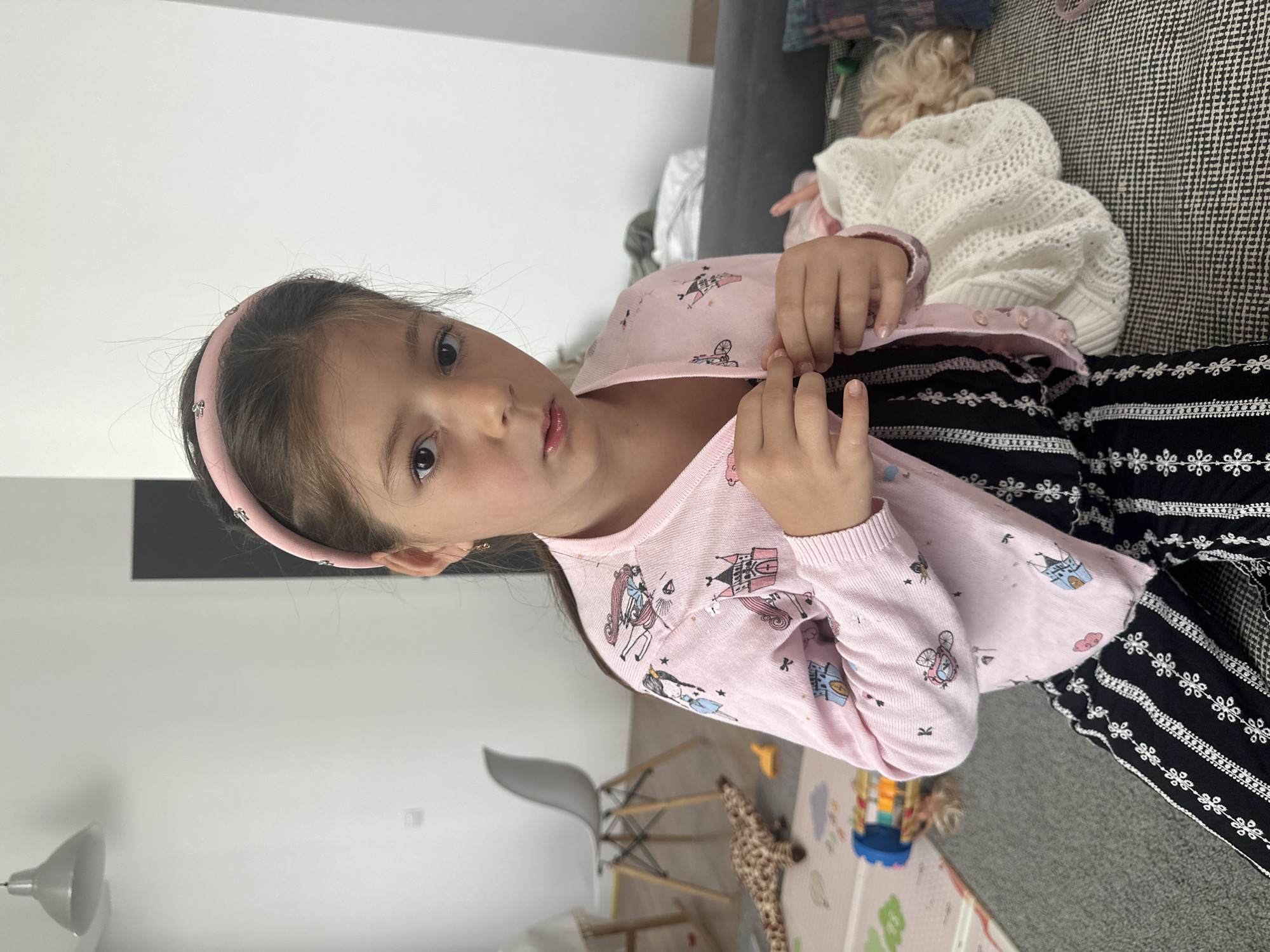 The media file shows a young girl wearing a pink cardigan with a fairy tale castle print and black and white striped pants. She is sitting on a couch, and her hands are holding open the front of her cardigan. Her gaze is directed towards the camera, and she has a pink headband on. Behind her, another child with blonde hair is visible, lying on the couch and covered with a white knitted blanket. The setting appears to be indoors, possibly a living room, with a toy giraffe and a floor mat visible in the background. The lighting suggests it is daytime.