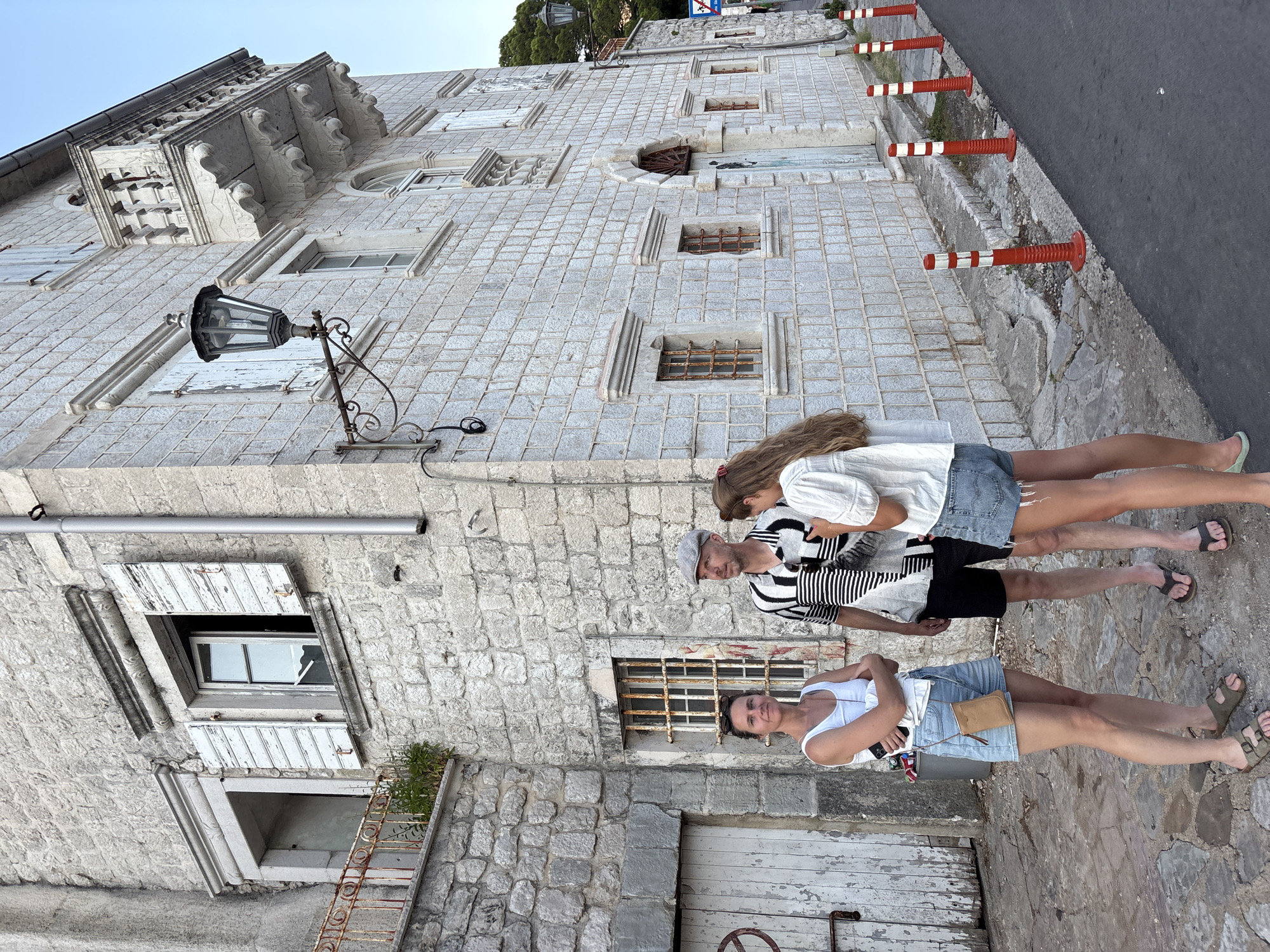 Three people, two women and one man, stand in front of a light-colored stone building in Perast, Montenegro. The building features weathered white shutters on some windows and a decorative balcony. The man is wearing a flat cap and a striped shirt, while the women are in casual summer attire. The ground is paved with uneven stones. Orange and white traffic cones line the adjacent asphalt road. The overall scene suggests an outdoor gathering on a street in a historic town.