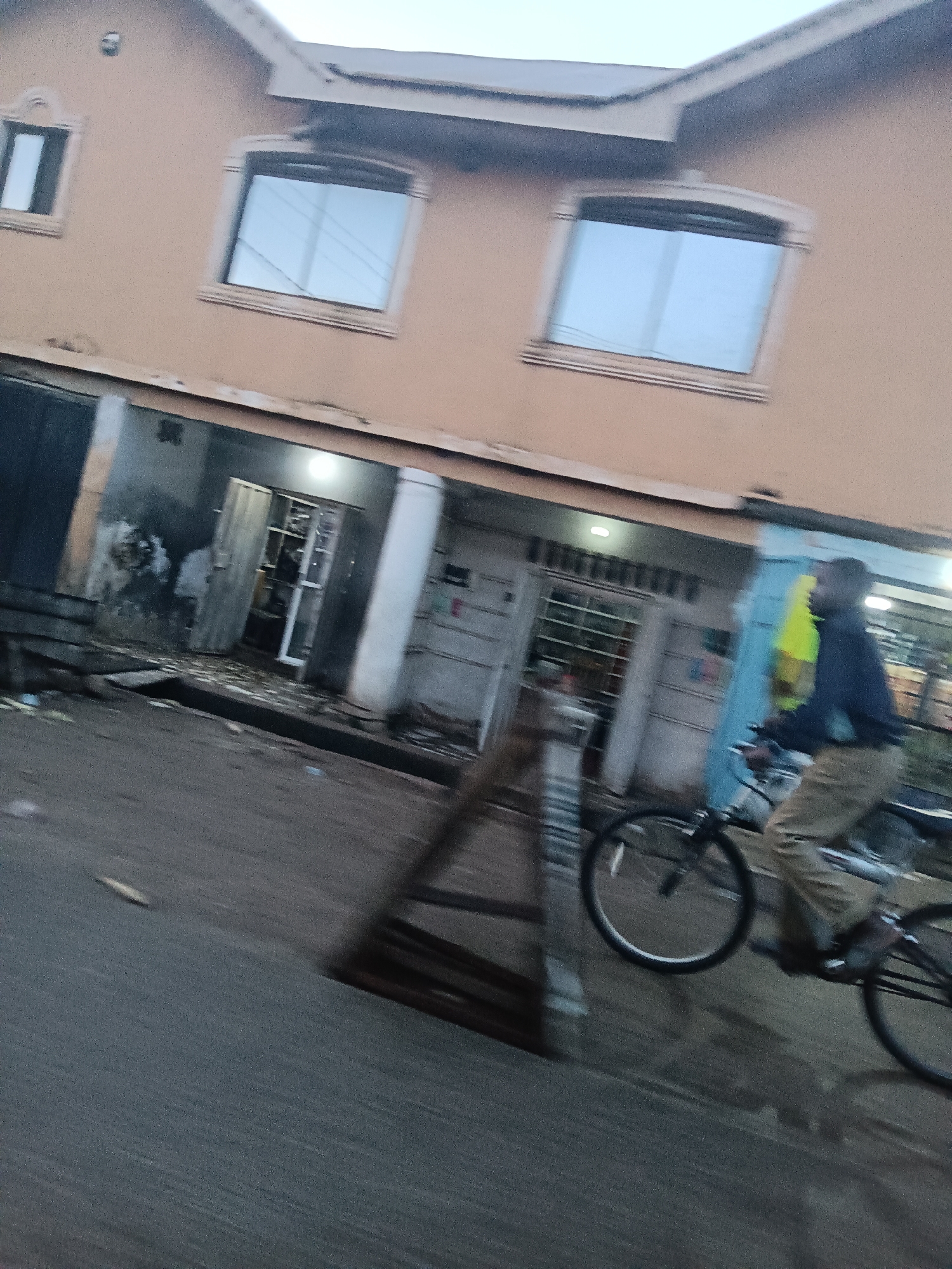 A man is riding a bicycle on a street in front of a two-story building with multiple windows and storefronts. The building is painted a light brown color. The man is wearing a dark jacket and light-colored pants. He is in motion, and the image is blurred due to the movement. The storefronts below the upper level appear to be open or have open doors. The street is paved and appears to be wet in some areas. There are some scattered debris on the ground in front of the storefronts. The sky is overcast. The scene suggests an urban environment.