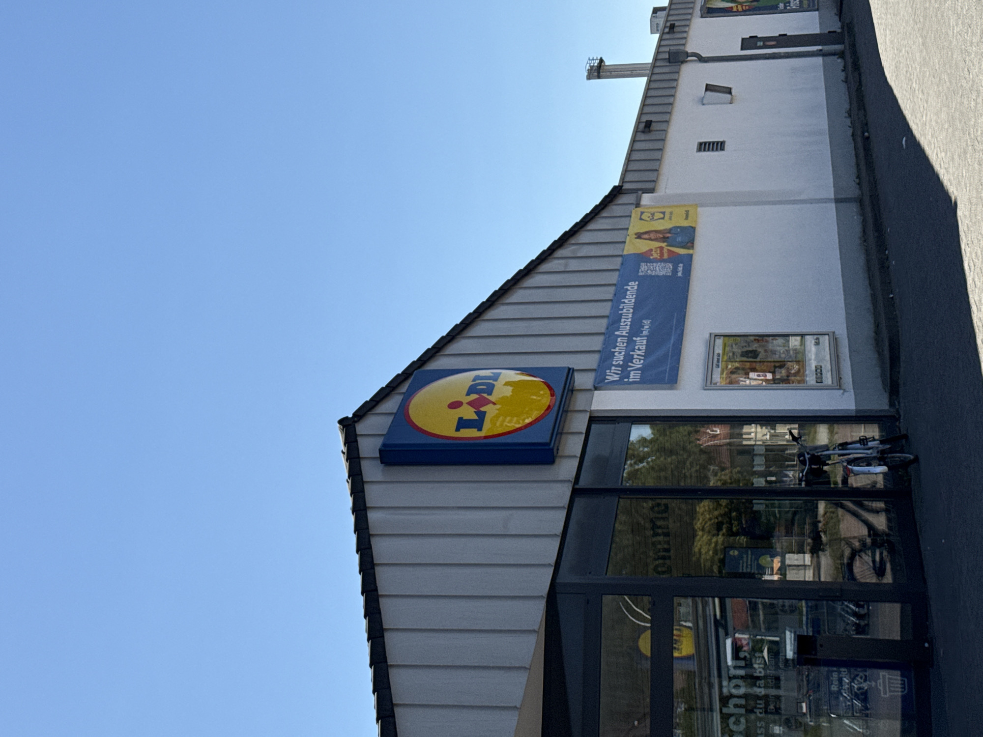 The image shows the exterior of a Lidl supermarket in Bad Lauterberg im Harz, Germany. The building has a grey, corrugated metal facade with a prominent blue and yellow Lidl logo above the entrance. To the right of the logo, a blue banner reads "Wir suchen Auszubildende im Verkauf (m/w/d)" which translates to "We are looking for trainees in sales (m/f/d)". There is a glass entrance with reflections of the interior and greenery outside. Two bicycles are parked in front of the store. The sky is clear and blue, suggesting a sunny day. The environment is urban, with a paved area in front of the store.