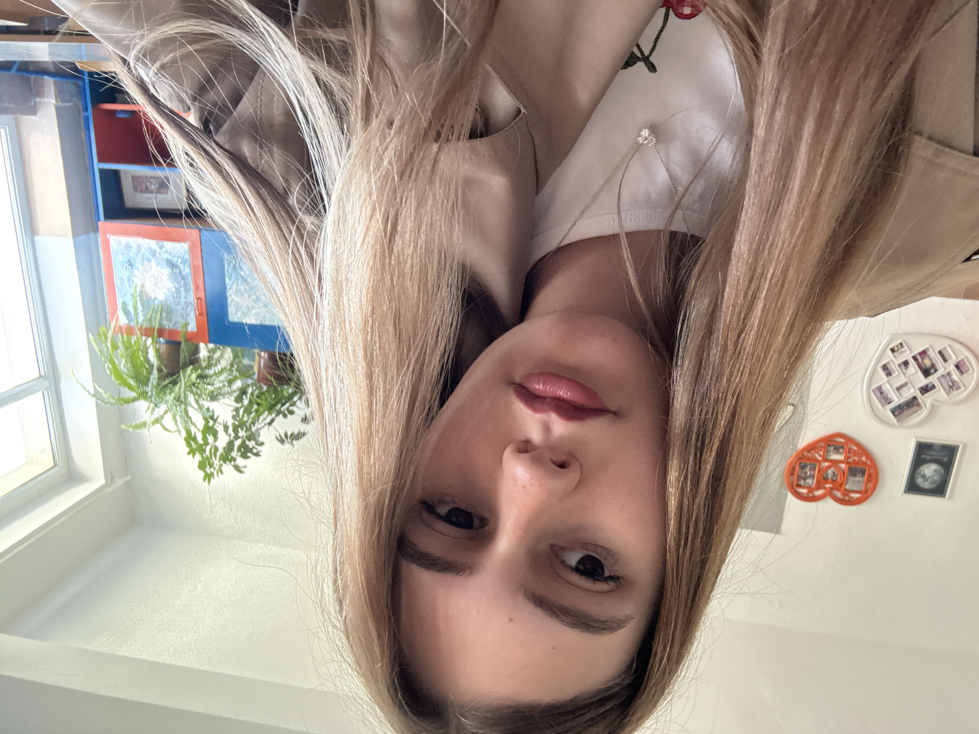 A close-up, selfie-style image of a young woman with light brown hair, wearing a beige trench coat and a white t-shirt with a cherry blossom design. The background includes a window with a plant, a blue and red cabinet, and a white wall with decorative photo frames. The location is in Panagyurishte, Bulgaria.