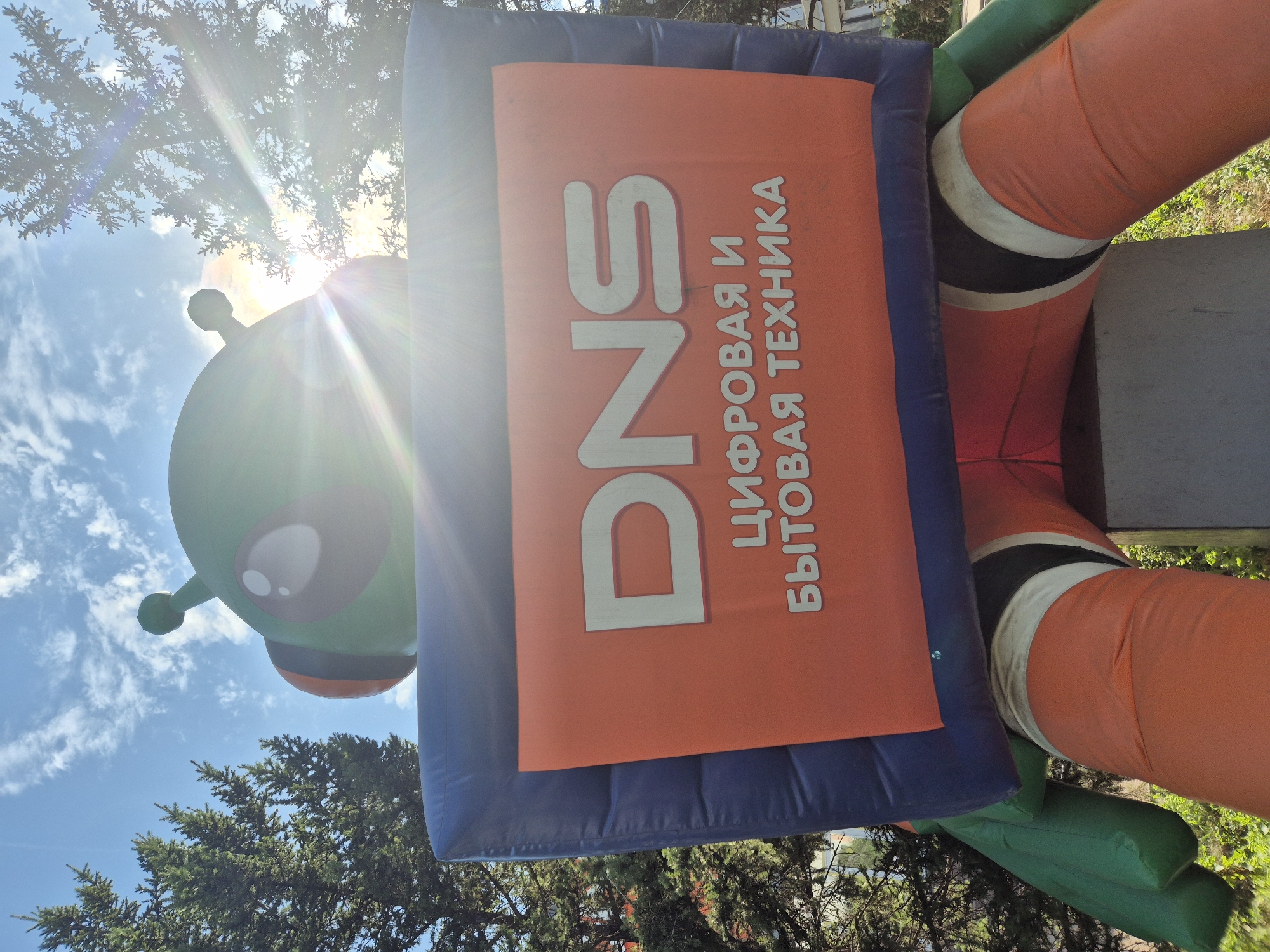 An inflatable alien mascot, green with large eyes and orange accents, sits with its legs spread. It holds a large rectangular sign with the DNS logo and the Russian text "ЦИФРОВАЯ И БЫТОВАЯ ТЕХНИКА" (Digital and Home Appliances) on an orange background, framed by a blue inflatable border. The mascot is positioned outdoors, with green trees and a blue sky with scattered clouds visible in the background. Sunlight flares brightly from the upper right of the image. This scene is located at a DNS store in Kokshetau, Kazakhstan.