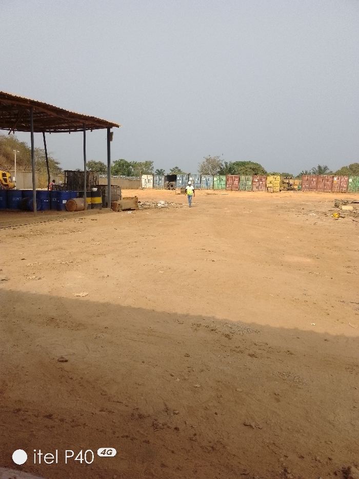 An outdoor industrial or storage yard in Hoche, Nigeria, is depicted under a hazy, light gray sky. The foreground and midground consist of a vast expanse of reddish-brown dirt, showing signs of vehicle traffic and scattered debris.
On the left side of the midground, a large, open-sided shelter with a corrugated metal roof is present, supported by dark poles. Beneath this shelter, several blue and yellow industrial barrels or drums are visible, some stacked and some lying horizontally. A partial view of yellow heavy machinery, possibly an excavator, is visible to the left behind the shelter. A person wearing a red shirt is partially visible under the shelter, near the barrels.
In the center of the midground, a person walks from left to right across the dirt. This individual is wearing a white hard hat, a yellow-green high-visibility vest over a light-colored shirt, and dark pants.
The background features an extensive line of various colored shipping containers (white, light blue, green, yellow, and rust/brown) arranged in rows across the entire width of the frame. Behind these containers and in the far distance, sparse vegetation and trees are visible. A fence is partially visible behind the shelter and some containers. A utility pole stands on the far left. The overall scene suggests a commercial or industrial storage operation.
