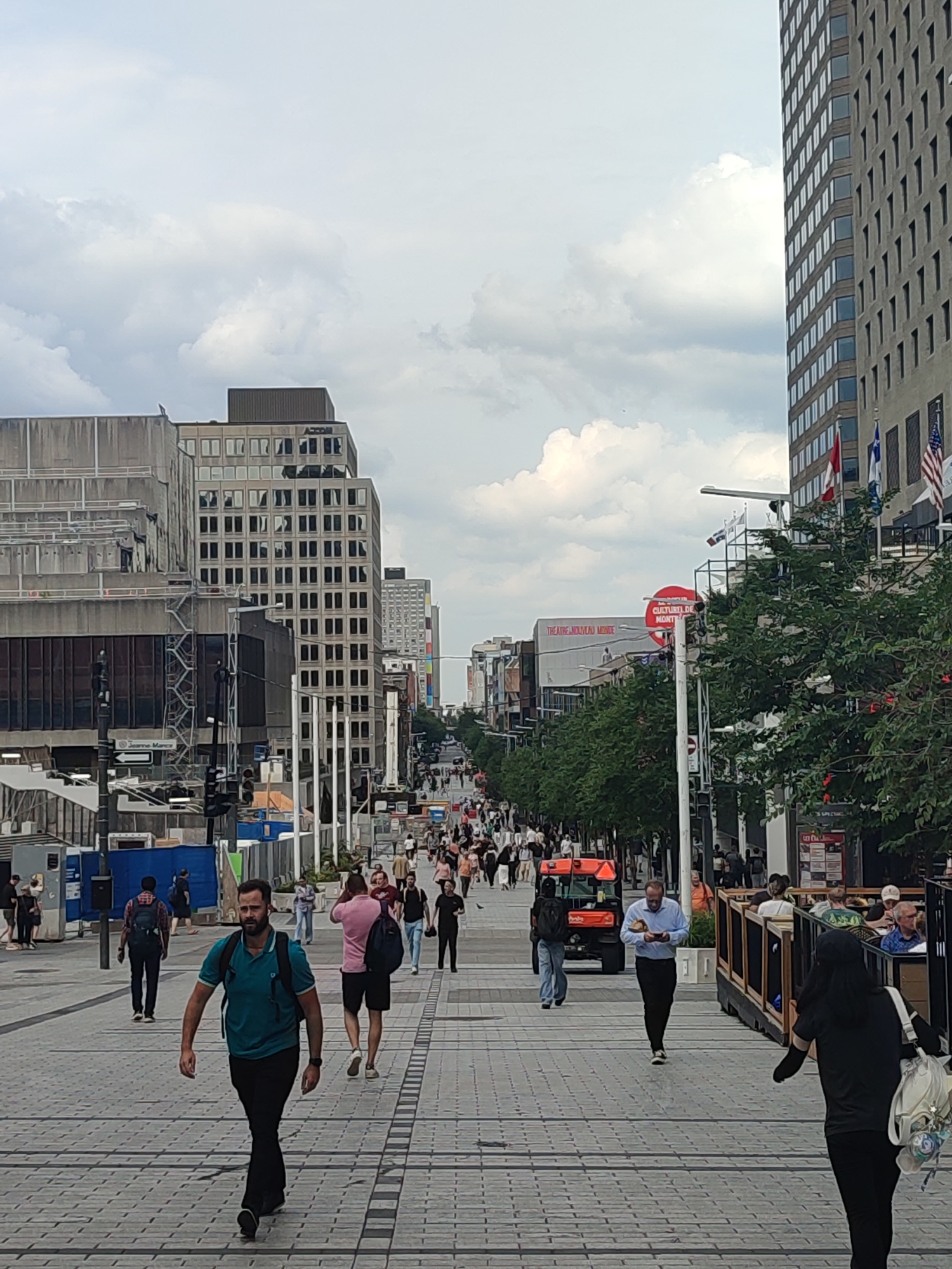The image depicts a busy pedestrian street in Montreal, Canada. The scene is captured from a slightly elevated perspective, looking down a wide, paved walkway lined with trees and buildings.
In the foreground, a man wearing a teal polo shirt and black pants walks towards the viewer. He has a backpack slung over his left shoulder. Further down the walkway, other pedestrians are visible, some walking in pairs and groups, others individually. They are dressed in casual clothing, suggesting a warm day.
The environment includes modern buildings with multiple stories and large windows. One building on the right has a distinctive architectural style with numerous vertical window elements. Trees are planted along the walkway, providing shade. Various signs and advertisements are visible on the buildings, including one that reads "THEATRE NOUVEAU MONDE" and "CULTUREL DE MONTRÉAL". A bright orange utility vehicle is parked on the street on the left side of the image. Flags, including the Canadian and Quebec flags, are displayed on poles.
The ground is paved with interlocking stone tiles in a grid pattern. The sky is overcast with scattered clouds. Construction or renovation appears to be taking place on the left side of the street, with barriers and scaffolding visible. The overall impression is a lively urban street scene on a cloudy day in Montreal.