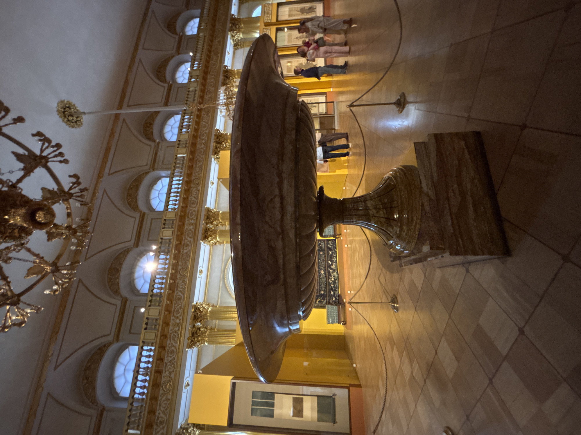 A wide-angle shot captures the interior of a grand, classical building, possibly a museum or palace, featuring ornate architecture and artistic exhibits. The scene is dominated by a large, sculpted marble vase that rests on a pedestal. The vase has a fluted design and a wide, shallow bowl. The surrounding space is adorned with golden architectural details, including intricate railings and columns, and arched windows that let in soft, diffused light. A large, elaborate chandelier hangs from the high ceiling.
In the background, several people are observing an exhibit on a wall, consisting of framed artwork. Their postures suggest casual observation and appreciation. The floor is tiled in a geometric pattern, reflecting the light and adding to the sense of spaciousness and grandeur. The overall impression is one of refined elegance and cultural display, likely during daylight hours given the illumination. The mood is tranquil and contemplative. No visible text is present in the image.
