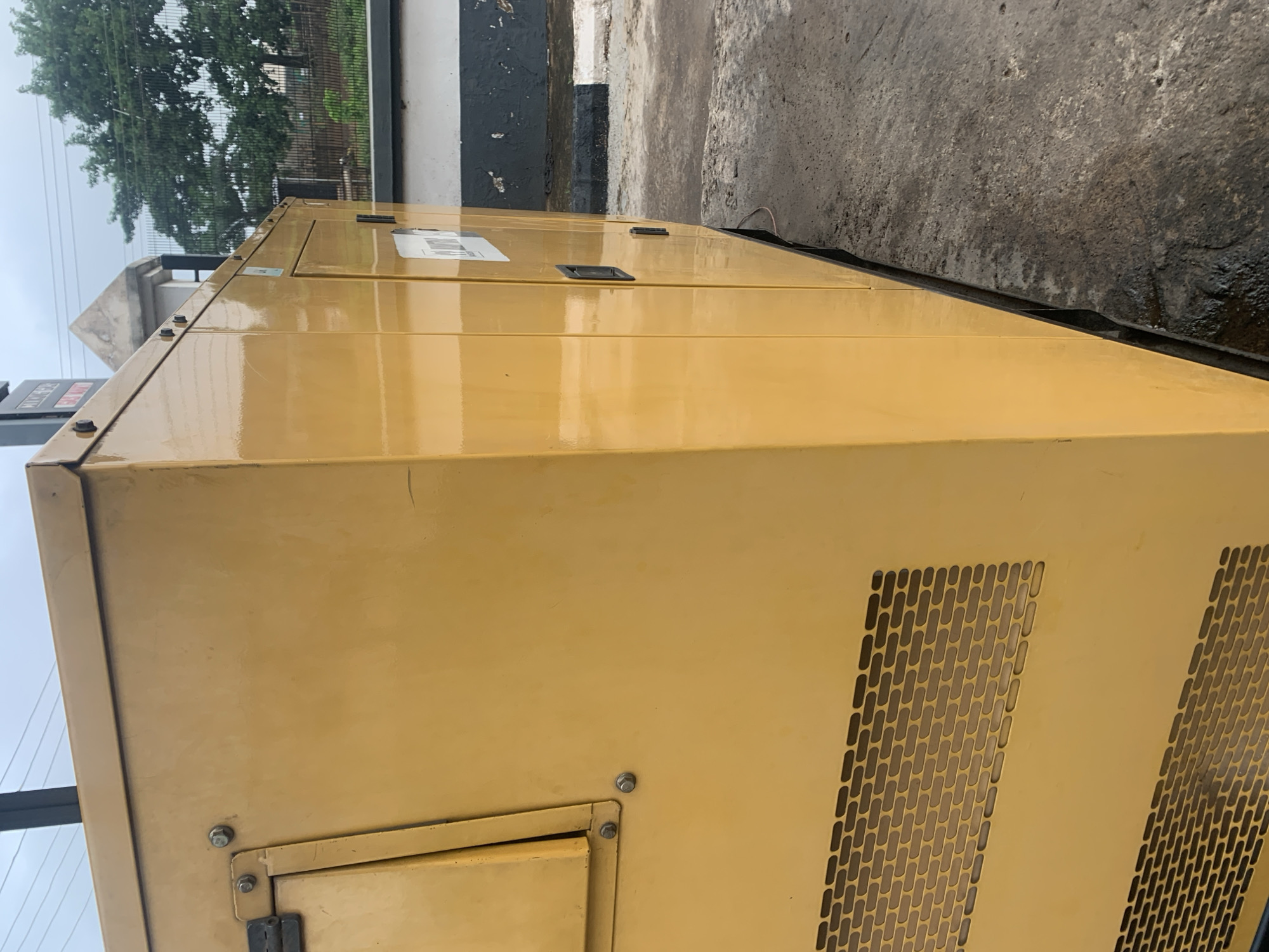 The image depicts a yellow generator enclosure in what appears to be an outdoor urban setting in Anilelerin, Nigeria. The generator is large and box-shaped, with visible ventilation grilles on its left side. A metal door with a black handle is present on its right side, with a smaller panel or latch to its right. There are visible bolts and seams on the enclosure, indicating its construction. The generator is situated on a concrete surface that shows signs of wear and some wetness. In the background, a black and white striped curb is visible, leading to a paved area. Beyond that, a low fence with green foliage and trees can be seen, suggesting a slightly vegetated area. The sky is overcast, indicating a cloudy day. There are no people or animals visible in the image. The scene has a functional, industrial feel, typical of an operational generator in a city environment.