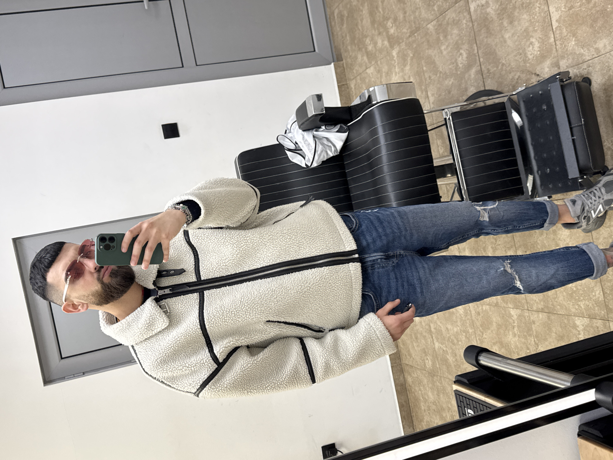 The image shows a man taking a selfie in what appears to be a barbershop. The man is wearing a white fleece jacket with black trim, blue jeans with rips, and gray sneakers. He is holding a smartphone in his left hand. He has short dark hair, a beard, and is wearing rose-tinted glasses.
Behind him, there is a barber chair and a mobile tool storage unit. The background consists of a white wall and a beige tiled floor. There is a door with a window in the wall to the left.
The location is Kazanlak, Bulgaria.