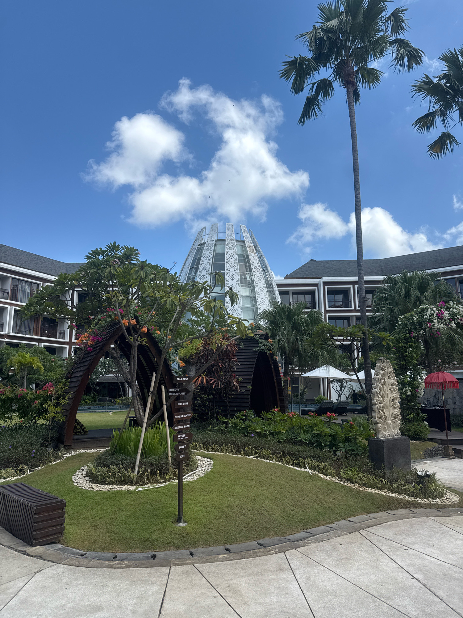 An eye-level, outdoor, medium shot captures a resort in Kuta, Indonesia, under a bright, partly cloudy blue sky. Palm trees dominate the upper right of the frame, with their fronds silhouetted against the sky. A distinctive modern building with a domed, white, latticework facade is visible in the background, partially obscured by lush green foliage and flowering trees. In the foreground, a wooden bench sits on a paved walkway. A circular garden with a manicured lawn, surrounded by white stones and various plants, features a directional signpost indicating "SPA & YOGA," "DHANYA RESTAURANT," "KIDS CLUB," "FITNESS CENTRE," "MEETING ROOM," "NAVEEN (BAR & GRILL)," "LOBBY," and "EXIT." A rustic wooden archway covered in greenery and flowers is positioned near the garden. In the mid-ground, lounge chairs and white umbrellas are visible near a swimming pool, and a carved stone sculpture stands to the right. A red umbrella and a table are also visible in the far right. The architecture of the resort appears to be a blend of modern and traditional styles. No people are visible in the scene.