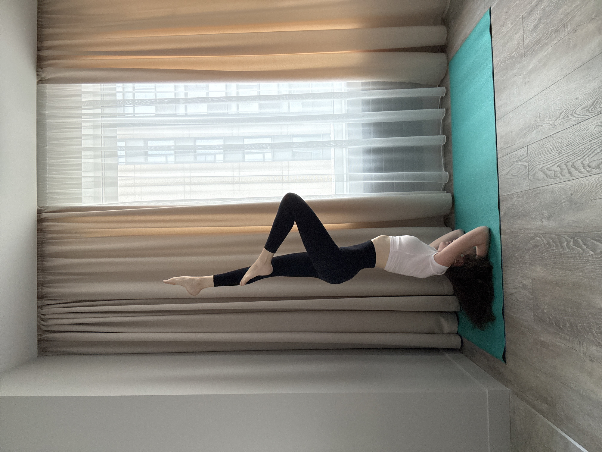 A woman is performing a yoga pose on a teal yoga mat on a wooden floor. She is in a headstand with her legs extended upwards. Her left leg is bent at the knee, and her right leg is straight. She is wearing a white t-shirt and black leggings. The background shows a window with beige and white curtains. The setting appears to be an indoor room. This image was taken in Moscow, Russia.