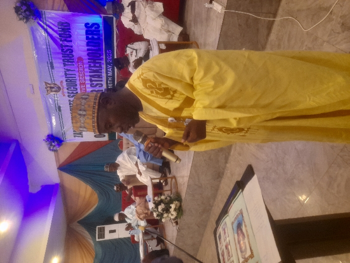 The image shows a man in a yellow robe speaking into a microphone. He is standing in front of a banner with the words "Zamfara State Security Trust Fund" on it. There are people seated in the background. The event is being held in a room with blue curtains and a large window. The image was taken in the city of Gusau, Nigeria, located at the coordinates 12.215, 6.676. The image shows a public event being held in Gusau, Nigeria. The event is about the Zamfara State Security Trust Fund and is being attended by many people. The man speaking into the microphone is likely a prominent figure involved with the security trust fund. The event is being held indoors in a space that is decorated with blue curtains and a large window. There is a table in the foreground that has a book on it, possibly related to the security trust fund. The image provides a glimpse into the efforts of the Zamfara State government in addressing security concerns in the region.