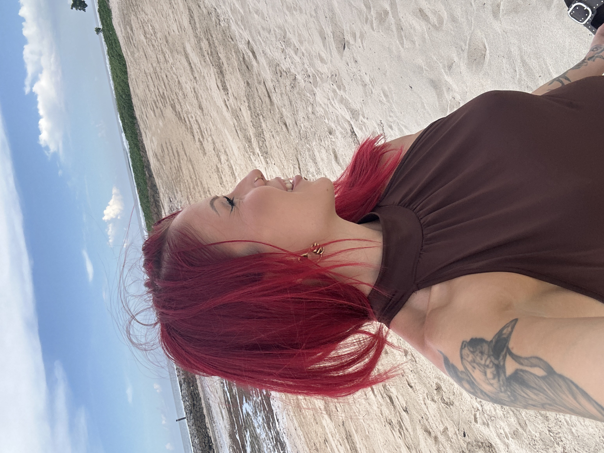 A woman with red hair and a brown dress is outdoors, seemingly on a beach, in Nusa Dua, Indonesia. She has a tattoo on her arm and is smiling and looking up towards the sky. The environment includes a sandy beach, the ocean, and the sky with clouds.