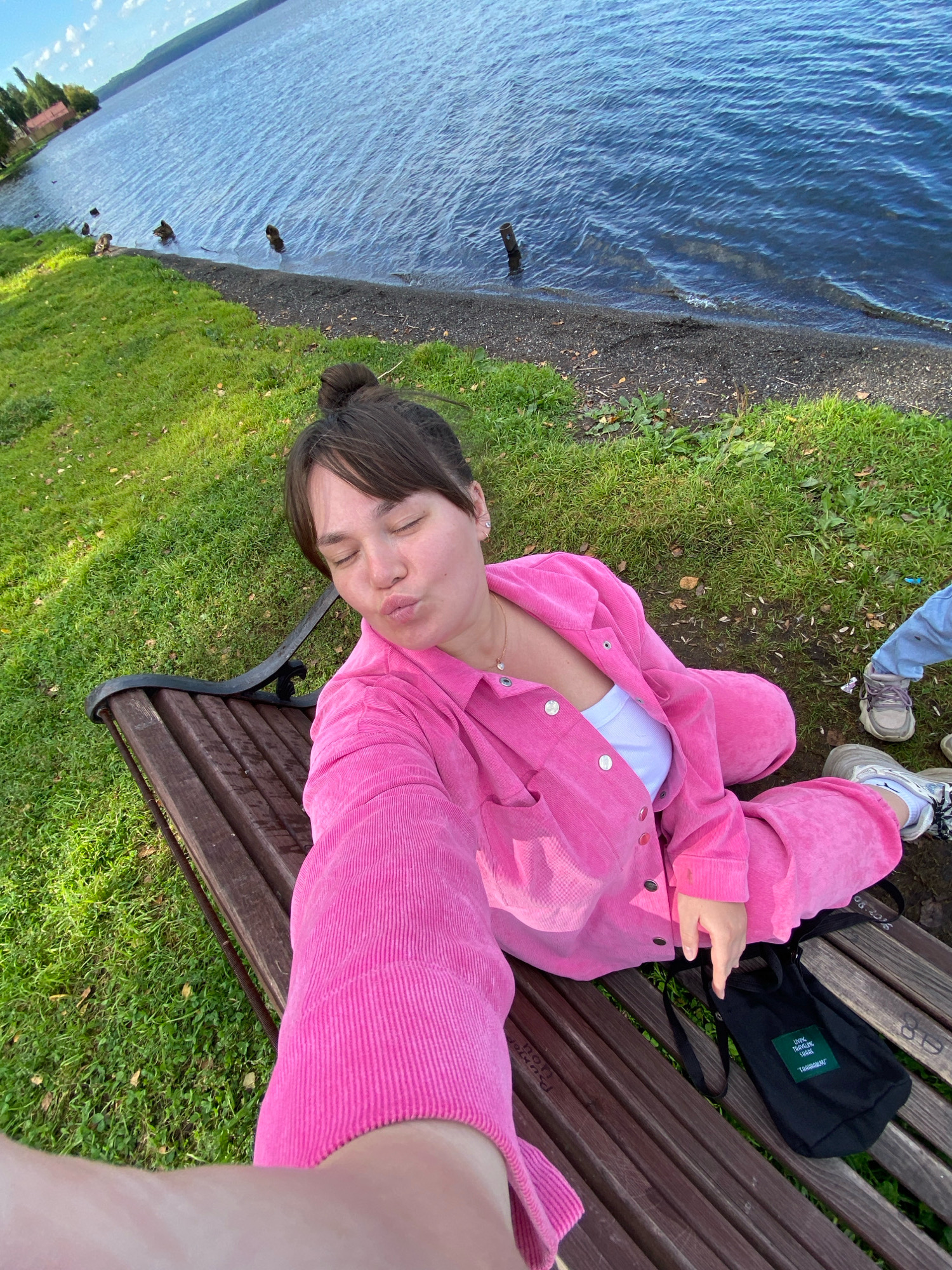 An outdoor, high-angle, selfie-style shot shows a young woman sitting on a wooden park bench next to a body of water. She is wearing a bright pink corduroy button-down shirt and matching pants, with a white tank top underneath. Her hair is pulled up in a bun, and she is puckering her lips as she takes the photo. Her eyes are closed. To her right, partially visible, are the legs of a child wearing light blue pants and sneakers. A small black canvas bag with green text rests on the bench to her right. The bench is situated on a grassy area with fallen leaves, adjacent to a dark gravel shoreline. Beyond the shore is a large body of blue water with gentle waves, and in the distance, a green hill with some trees and buildings. The overall setting appears to be a park or waterfront area. The presence of the water and the hilly landscape is consistent with the topography of Votkinsk, Russia, which is situated on the shore of Lake Votkinskoye.