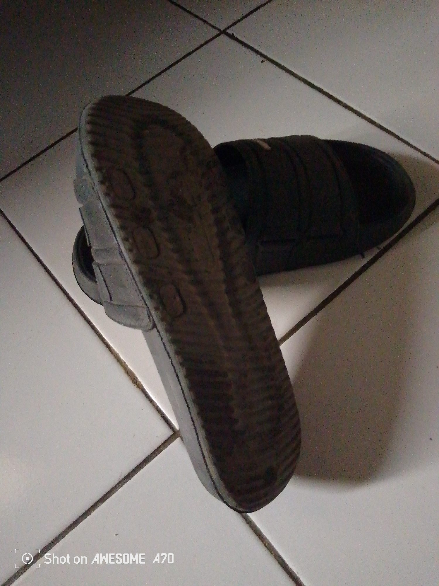 This is a close-up, high-angle shot of a pair of dark-colored flip-flops on a tiled floor. The flip-flops are positioned with one sole facing upwards and slightly to the left, revealing its worn, ribbed tread pattern and a textured footbed. The other flip-flop is partially visible behind and to the right of the first, with its upper strap facing upwards. The tiled floor appears to be composed of square tiles with dark grout lines, creating a grid pattern. The lighting is dim, casting a shadow from the flip-flops onto the floor. The bottom left corner of the image contains a watermark that reads "Shot on AWESOME A70". The image is taken in Cisaat, Indonesia.