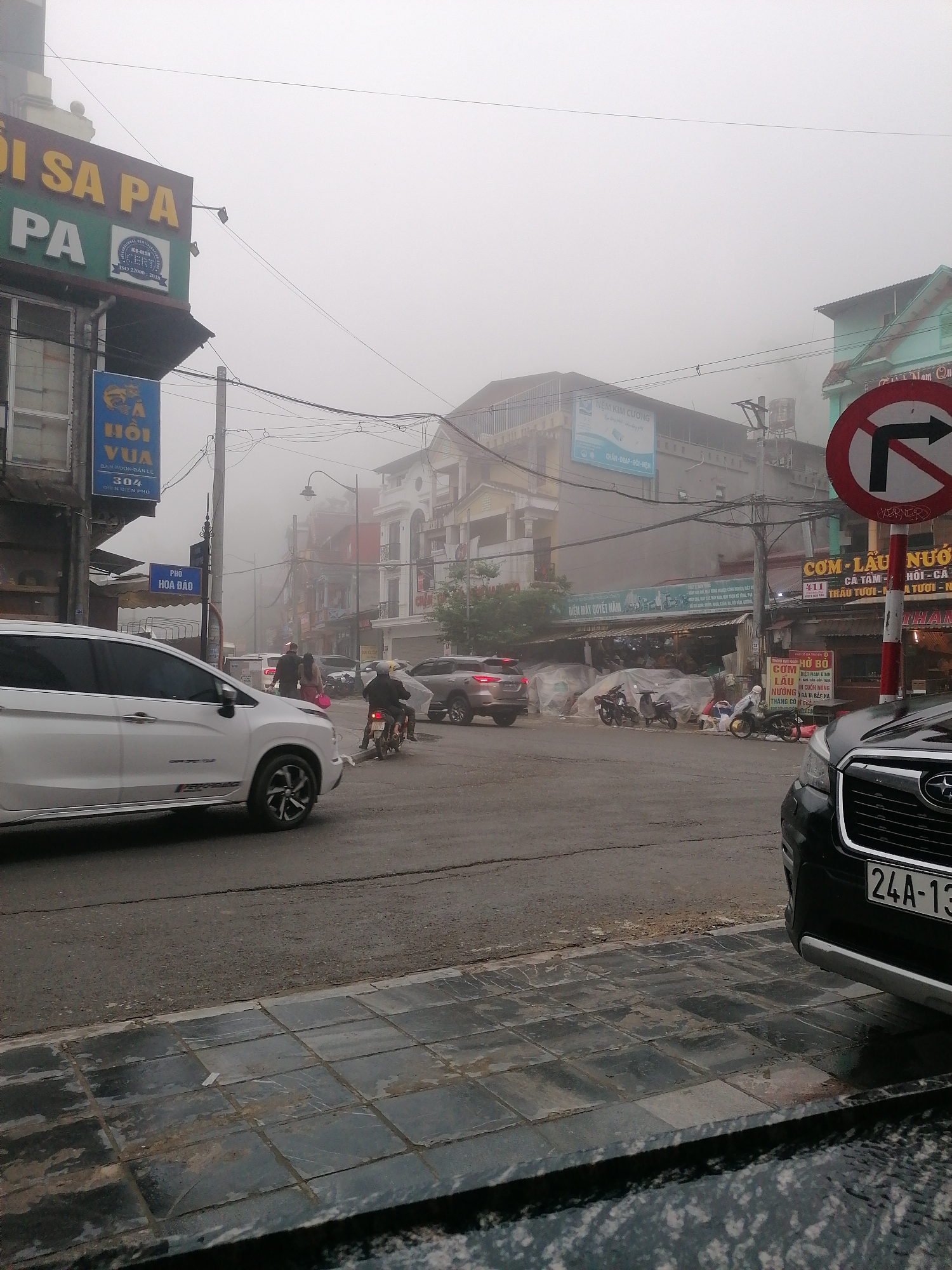 The image captures a bustling street scene in a town, shrouded in dense fog or mist, giving the environment a soft, diffused light and reduced visibility. The ground appears wet, reflecting the damp conditions.
In the foreground, a white SUV is prominent on the left, turning, while a black SUV is partially visible on the right, also at the curb. The license plate of the black SUV, "24A-13," is clearly seen. Several other vehicles, including a grey SUV and multiple motorcycles or scooters, are scattered across the intersection and parked near storefronts. People are visible on foot and riding scooters, engaged in daily activities.
The setting is an urban intersection lined with multi-story buildings that house various shops and businesses. Overhead, a tangle of power lines stretches across the street. A red circular traffic sign indicating "No right turn" is prominent on the right side of the image.
Visible text on signs further defines the location and commerce. A large sign on the left reads "SA PA," with a certification logo "ISO 22000:2018." Below it, a blue sign advertises "HỒI VUA" with "304 ĐIỆN BIÊN PHỦ" (Dien Bien Phu), a common street name in Vietnam. Other shop signs display "NỆM KIM CƯƠNG" (Kim Cuong Mattress), "PHỐ HOA ĐẢO" (Hoa Dao Street), and various food-related text like "CƠM LẨU NƯỚC," "CÁ TẦM - HỒI," "THỊT BÒ," and details about different dishes, suggesting eateries. The heavy fog is the most striking atmospheric detail, lending a mysterious and cool ambiance to the otherwise busy street.