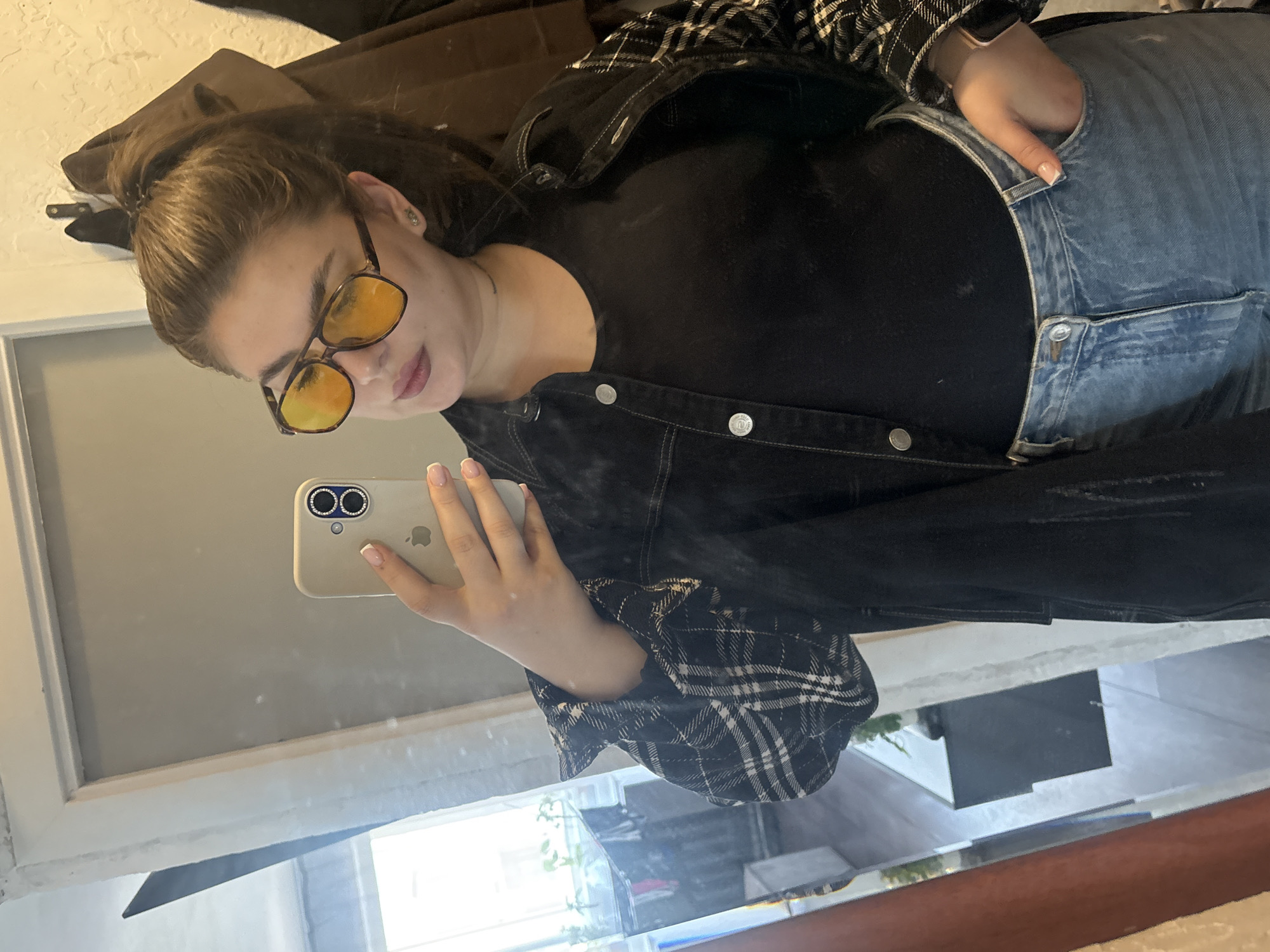 A woman is taking a selfie in a mirror. She is wearing yellow sunglasses, a black top, a black and white plaid jacket, and blue jeans. Her right hand is in her jeans pocket, and she is holding a phone with her left hand. The background is blurry, but it appears to be a room with a window. There is no evidence of this location being in the city of Chaikovsky, Russia.