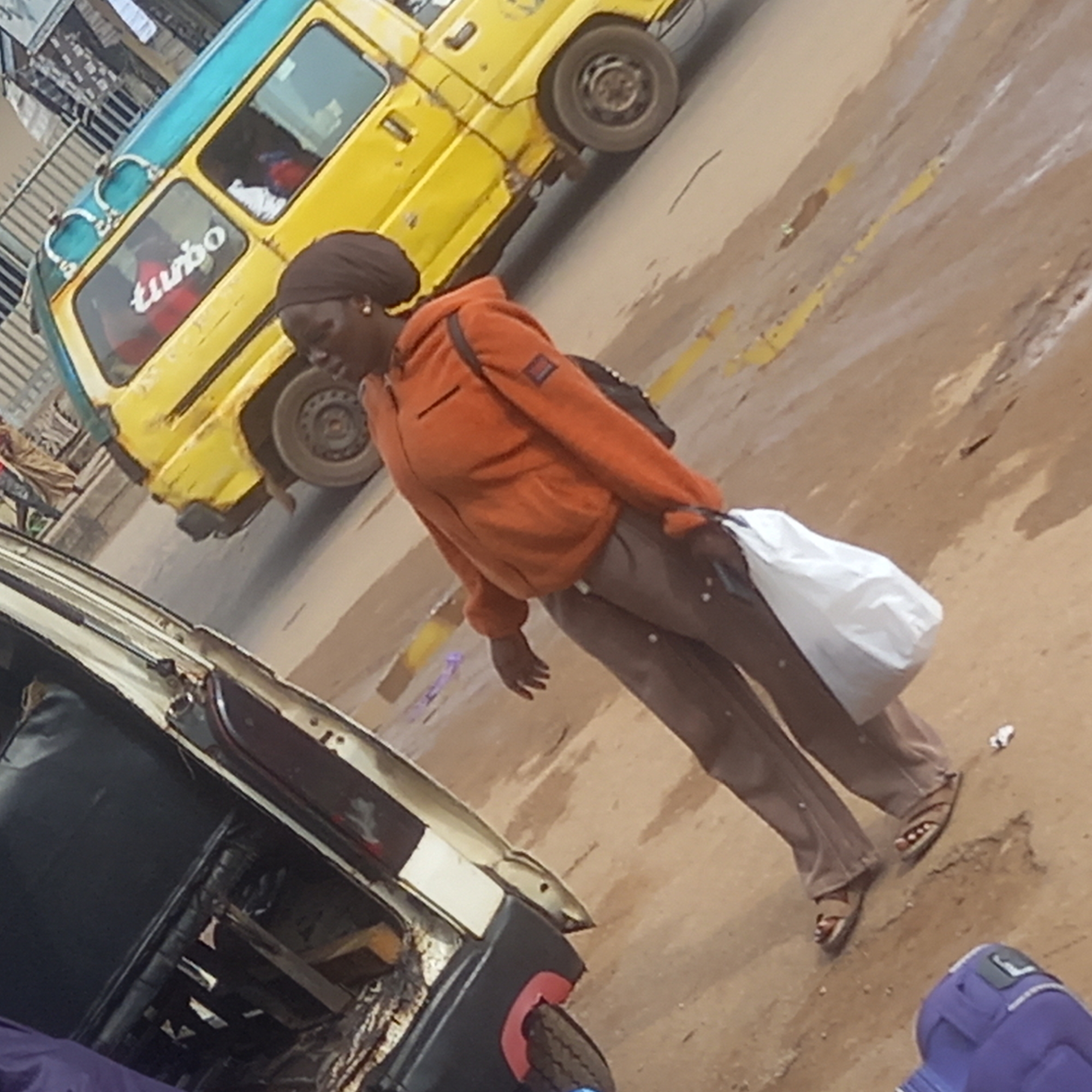 A woman stands on a street in what appears to be Sardauna, Nigeria. She wears an orange jacket, brown pants, and sandals. She carries a white plastic bag. Behind her is a yellow and blue vehicle with the word "Turbo" visible on the side. To the left, partially obscured, is another vehicle. The street surface appears to be a combination of dirt and paved areas, with some debris scattered around. The environment suggests an urban setting.