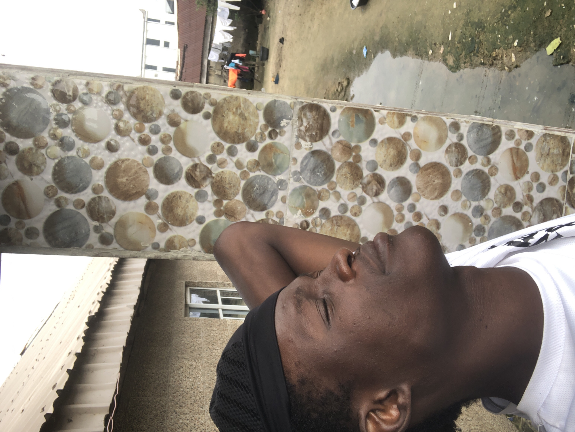 The image shows a young man in Ado, Nigeria, looking up with his eyes closed, his left arm bent behind his head. He is wearing a black cap and a white t-shirt. He is standing next to a pillar decorated with a mosaic of circular tiles in various shades of brown, grey, and white.
In the background, a beige building with a corrugated metal roof is visible. A small window with white panes is set into the building's wall. Further in the background, there is a courtyard with a muddy ground and some puddles of water. Clothes are hanging on a line between two poles. Two-story buildings with windows and a reddish-brown roof are also visible in the distance. The sky appears overcast.