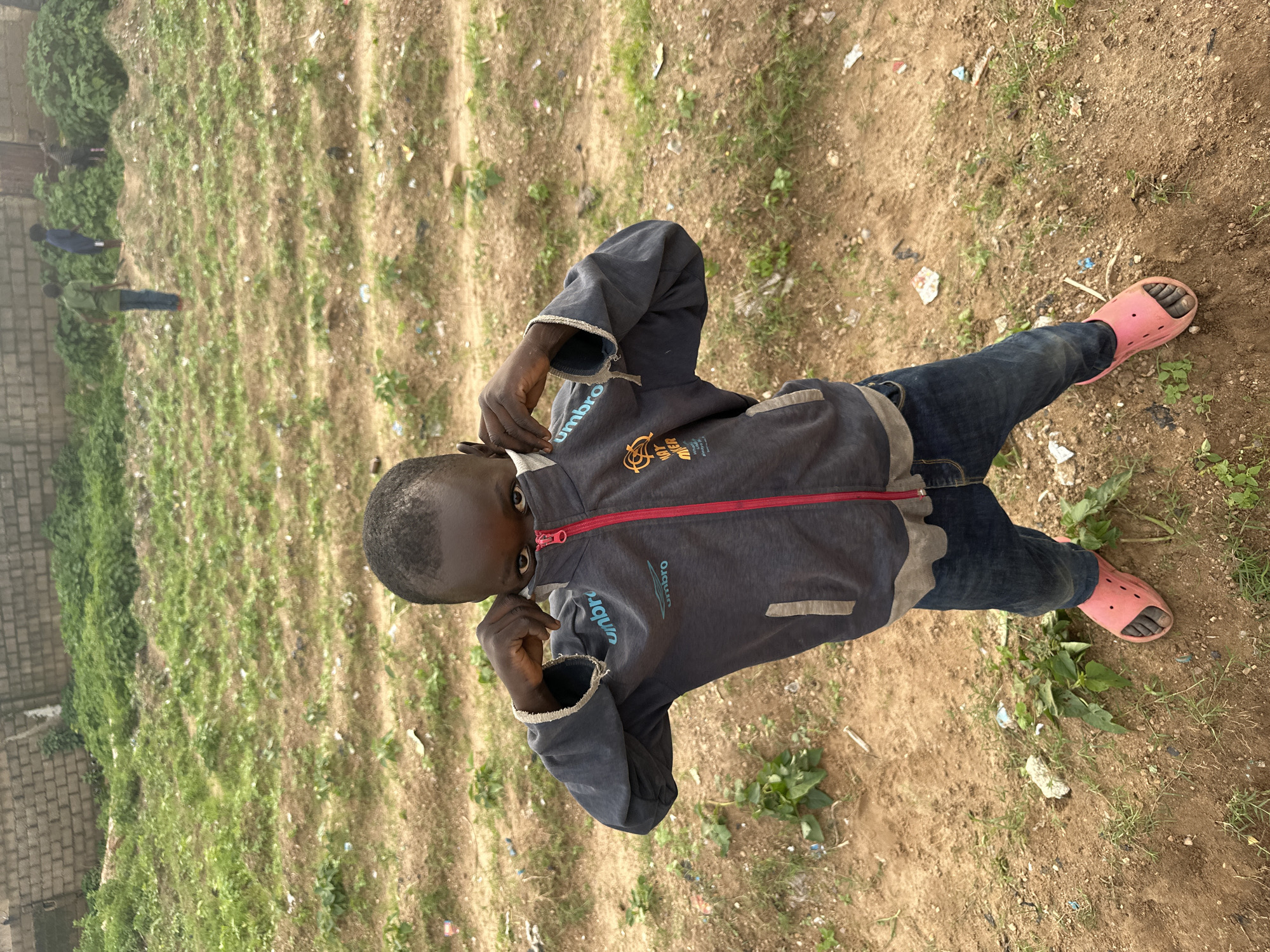 A young boy stands in a field of sparse vegetation and dirt on Rukuba Road, Nigeria. He is wearing a dark grey zippered jacket with the "umbro" logo visible on the chest, blue jeans, and pink slides. The boy is pulling the collar of his jacket up to cover his mouth and nose, looking directly at the camera. In the background, other children are visible playing on a grassy hill, and a brick wall borders the area. The ground is uneven with patches of dry earth and small green plants, littered with some debris.