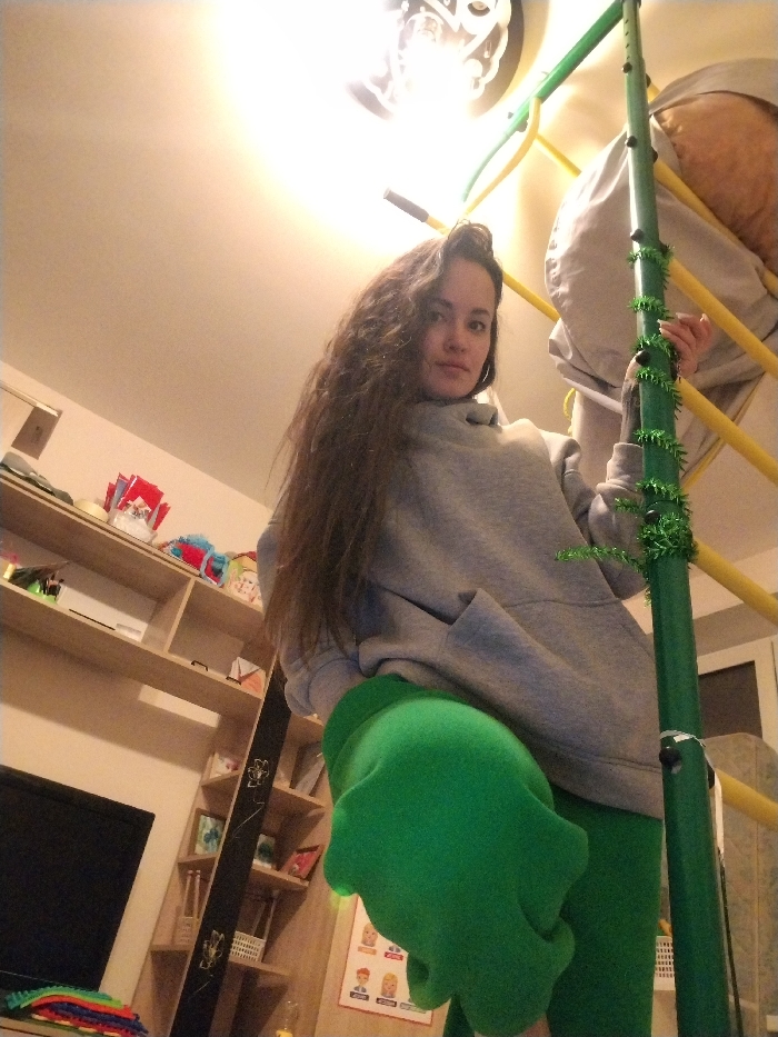 The image depicts an indoor residential scene in the city of Noviy, Russia. A female subject is prominently featured, positioned to the right of the frame, looking directly at the camera. She has long, dark, wavy hair and is wearing a grey hooded sweatshirt with a large front pocket and bright green pants. Her right hand grips a vertical green pole that forms part of an indoor play structure. This pole is wrapped with green tinsel-like garland and features some dark spherical attachments. Behind the subject and to her right, a light grey fabric-covered object, possibly a padded element, hangs from yellow horizontal bars, which are also part of the play structure.
To the left of the frame, a wooden shelving unit stands against a light-colored wall. The shelves hold various items, including red and white bags or boxes, one with visible "Marlboro" branding, and other colorful objects. Below the shelving unit, a dark flat-screen television is visible on a surface, in front of which a stack of colorful textile cloths is placed. A tall, thin, dark object with light-colored decorative elements stands between the TV and the shelves. On the wall further to the right, a poster or chart displaying illustrations of multiple human heads is partially visible. The ceiling is light-colored, with a bright, exposed light fixture illuminating the upper central portion of the image.