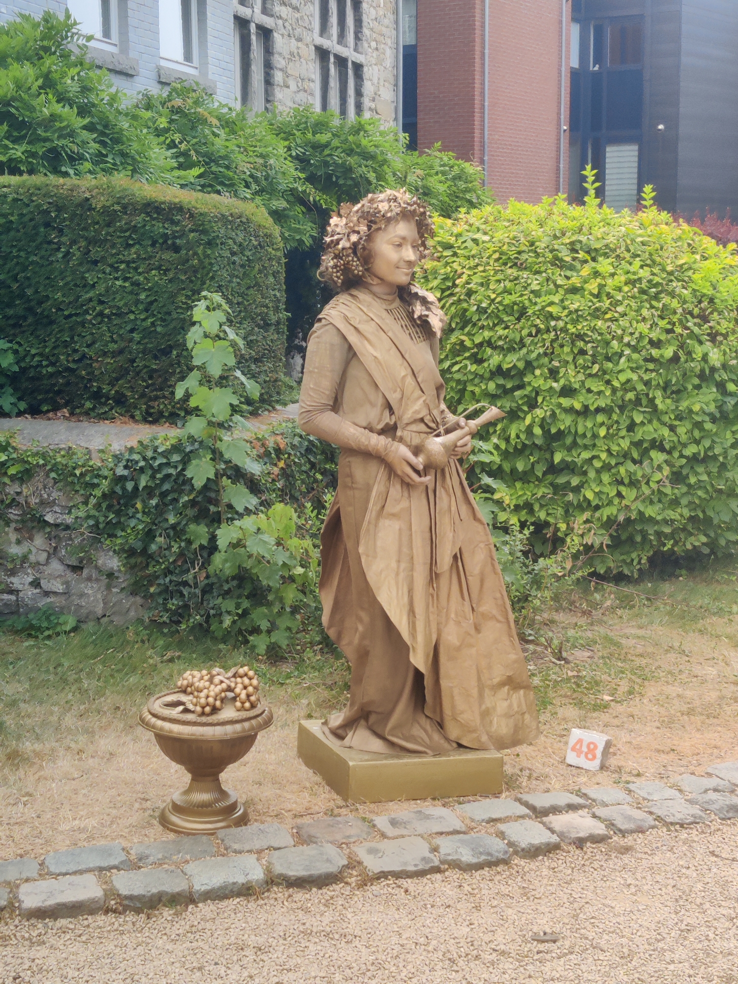 A living statue, its body and costume painted a uniform gold, stands in an outdoor setting, likely a public park or garden. The statue depicts a figure wearing a floral wreath on its head and a draped garment. It holds a small, ornate jug in its hands. In front of the statue, to its left, is a golden urn filled with what appears to be decorative grapes. A small stone cube with the number "48" in orange is visible to the right of the statue's base. The background features lush green hedges, ivy-covered stone walls, and a stone building, suggesting a historical or scenic location. The ground is covered with small gravel and paved with flagstones. The statue is positioned near a path made of these flagstones. The overall impression is that of a street performer situated in a public space. The context of Marche-en-Famenne, Belgium, implies this is a scene from that specific city.