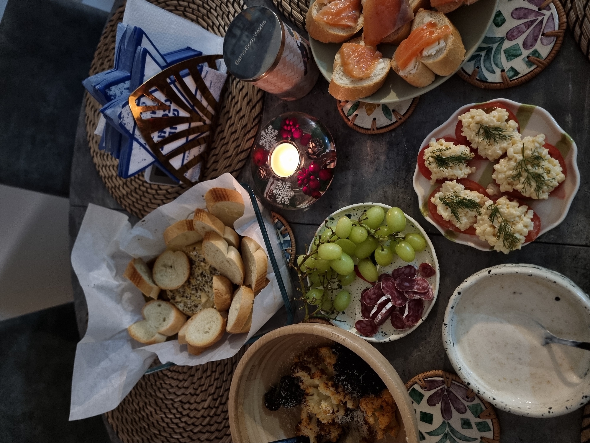 The image depicts a spread of food items arranged on a dark, possibly wooden, surface. There is a wicker tray holding blue and white napkins in a gold-colored holder, a Bath & Body Works candle, and a bowl of sliced bread. A small, decorative candle holder with a lit candle sits near the bread. Additionally, there is a plate of open-faced sandwiches with salmon, a plate of tomato slices topped with a white mixture and dill sprigs, a plate of green grapes and sliced salami, and a wooden bowl containing food with dark toppings. A white bowl with a spoon inside is also present. The location, Ramat Gan, Israel, is not discernible from the visual elements in the image.