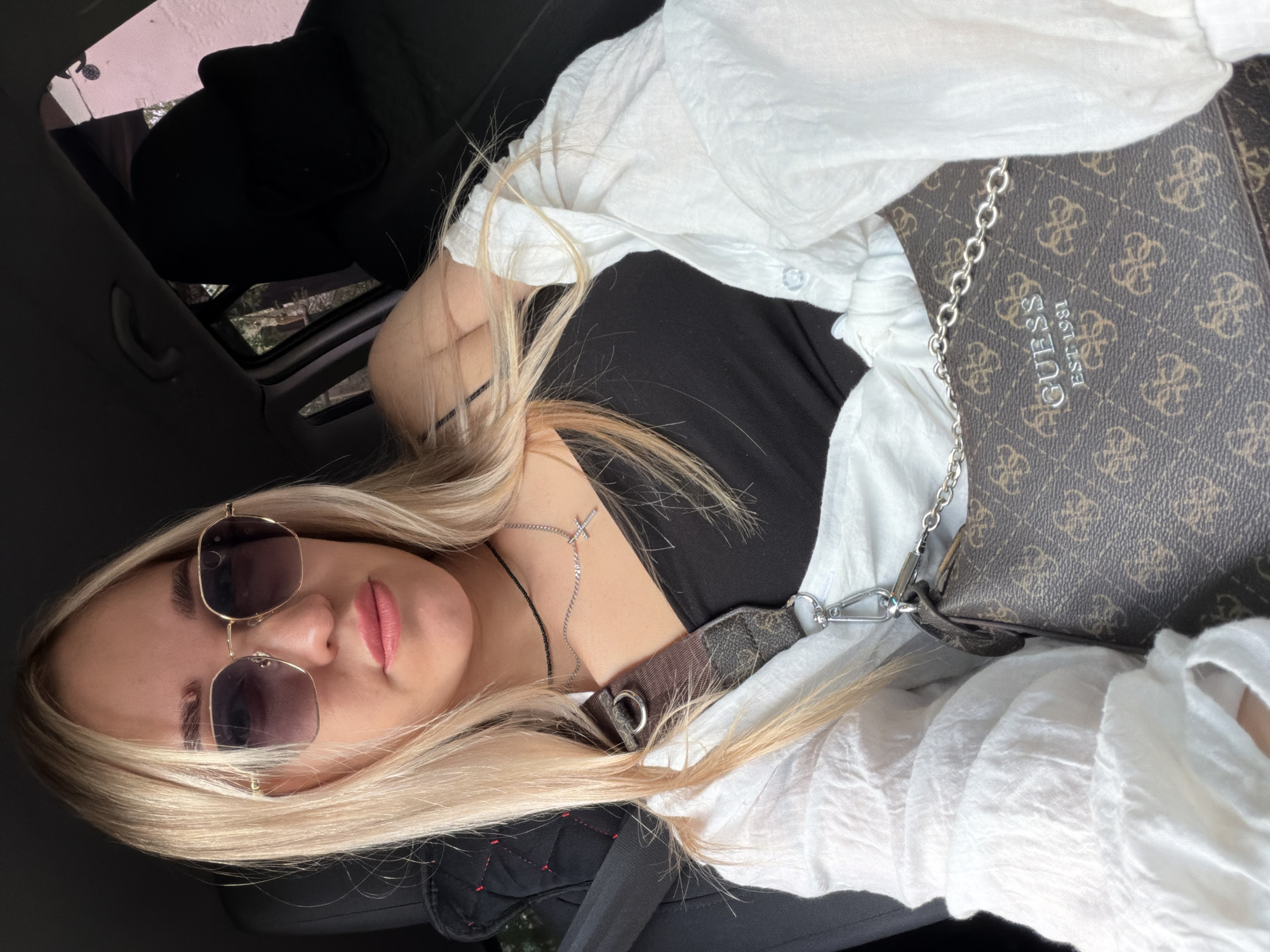 A blonde woman wearing sunglasses, a black tank top, and a white button-up shirt is seated in a car. She has a Guess purse with a chain strap. The car's sunroof is open. The location is Unknown, Unknown.