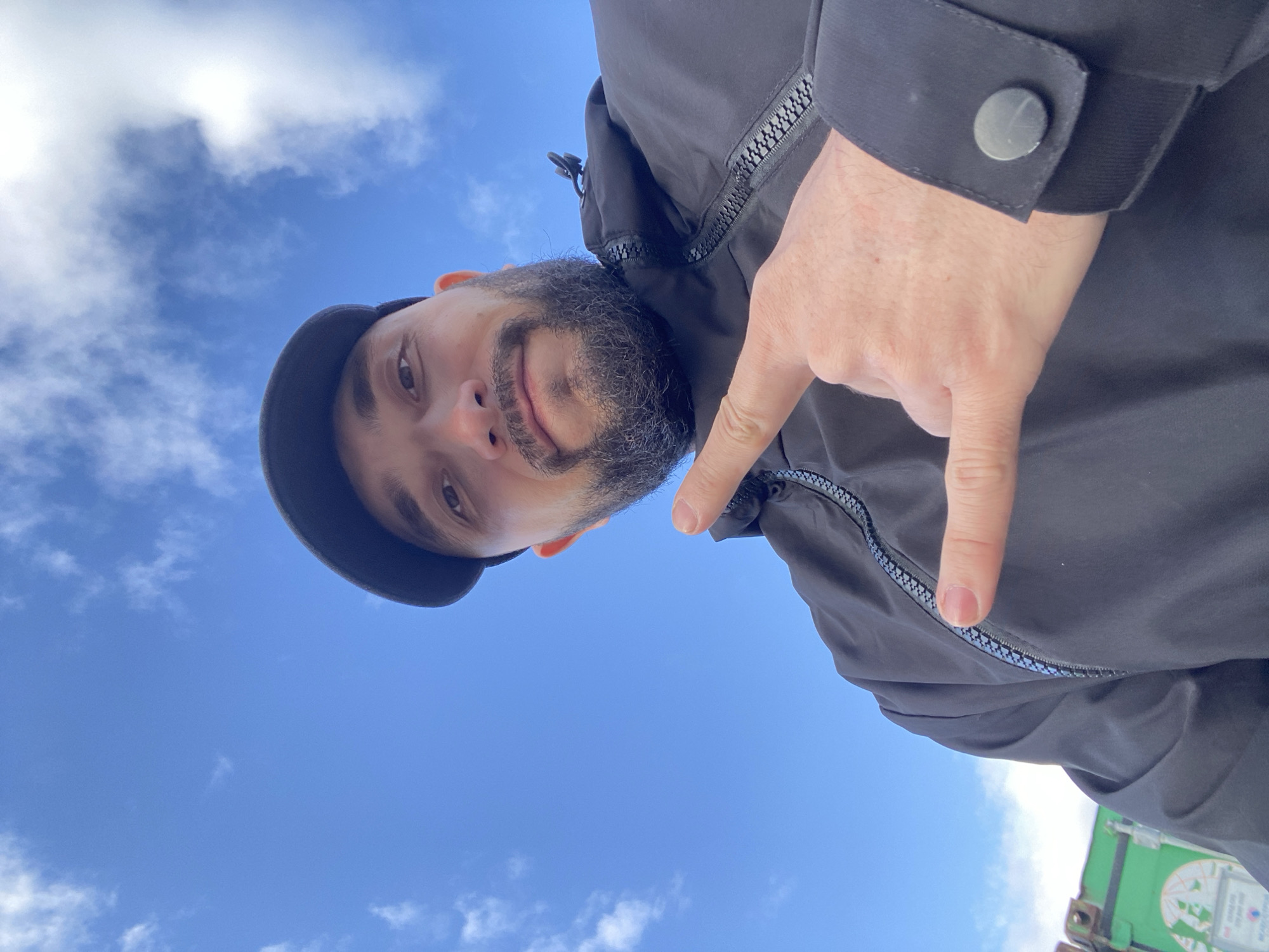 The image is a low-angle selfie of a man wearing a black cap and jacket. The man has a beard and is pointing his finger down towards the lower right corner of the frame. The background consists of a blue sky with white clouds. A small portion of a green object is visible in the very bottom right corner. The image location cannot be determined based on the image.