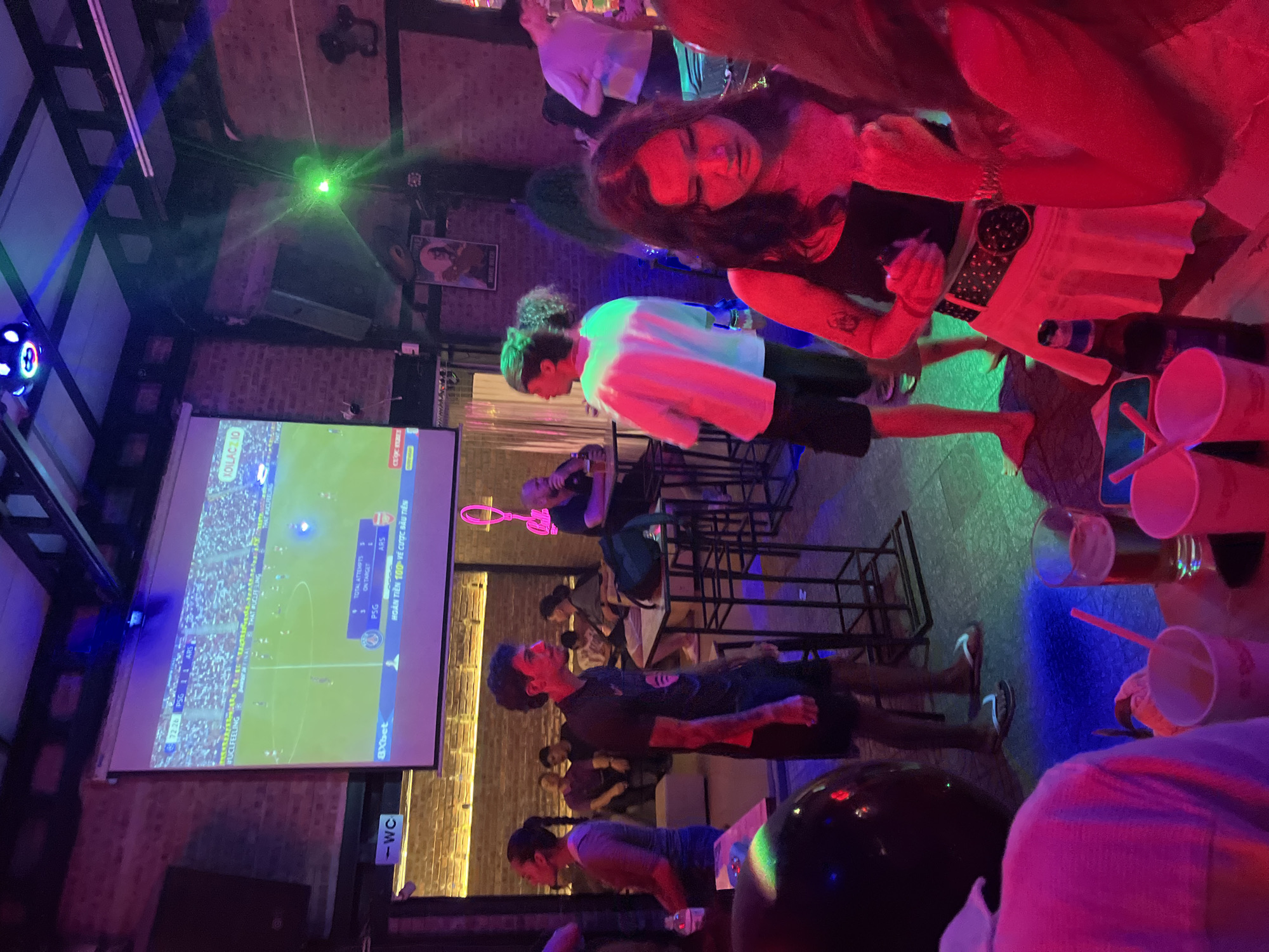 The image depicts a lively scene inside a bar in Đà Nẵng, Vietnam. People stand and sit, watching a soccer game projected on a large screen displaying the score as PSG 1 and ARS 1. The bar is dimly lit with blue, green, and red lights. A woman with long dark hair stands in the foreground, facing the right side of the image. Several plastic cups and a beer bottle sit on a nearby table. Various patrons are scattered throughout the establishment, some standing and others sitting on bar stools.