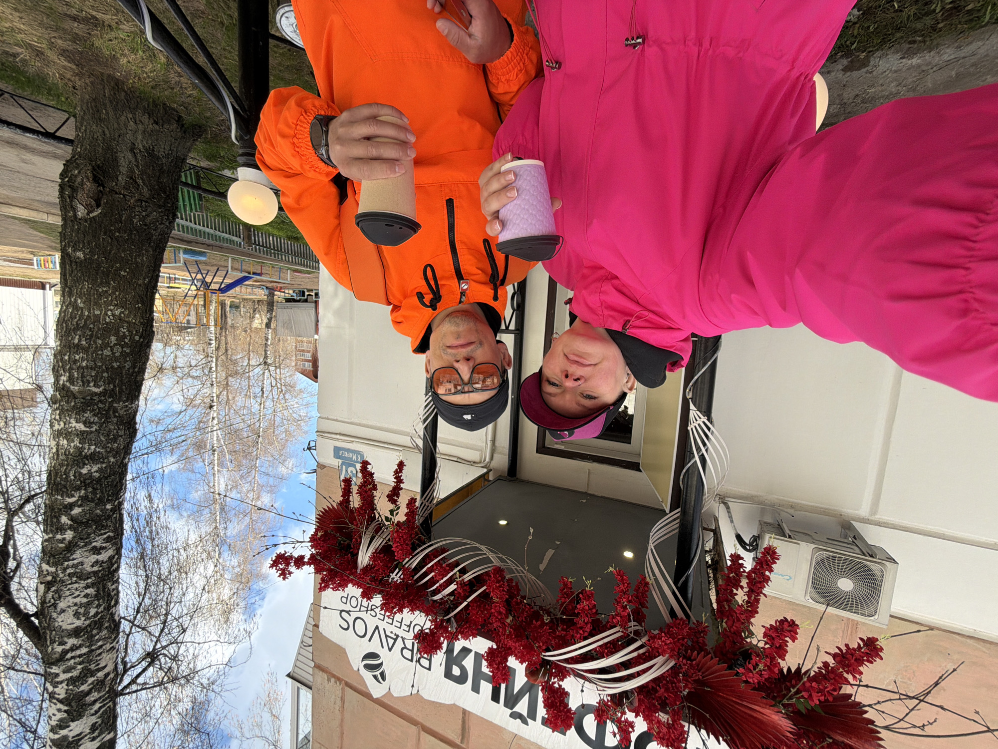 The image shows two people, a man and a woman, posing outdoors in front of a building, likely in Chaikovsky, Russia. The man, on the left, wears an orange anorak jacket, sunglasses, and a black baseball cap. He holds a light-colored reusable coffee cup. The woman, on the right, wears a bright pink anorak jacket and a dark pink baseball cap, holding a purple textured reusable coffee cup. Both are looking directly at the camera with neutral to slightly smiling expressions.
The scene is set during daytime, under a clear sky with some clouds. To the left, a large tree with bare branches, possibly a birch, suggests late winter or early spring. In the background, there's a children's playground with colorful equipment.
The building's facade is adorned with striking red floral or leafy decorations and white ribbons, especially above the entrance. An outdoor air conditioning unit is visible on the building. A street lamp with an illuminated bulb is also present near the man.
Visible text includes "PRAVOS COFFEESHOP" and a coffee bean logo on the building's sign. Below it, in larger Cyrillic letters, "ПИВНОЙ" (Pivnoy, meaning "Beer") is partially visible, suggesting a combined establishment or adjacent businesses. On a blue street sign on the corner of the building, "УЛ. К. МАРКСА" (Ul. K. Marksa – Karl Marx Street) and the number "31" are clearly legible.