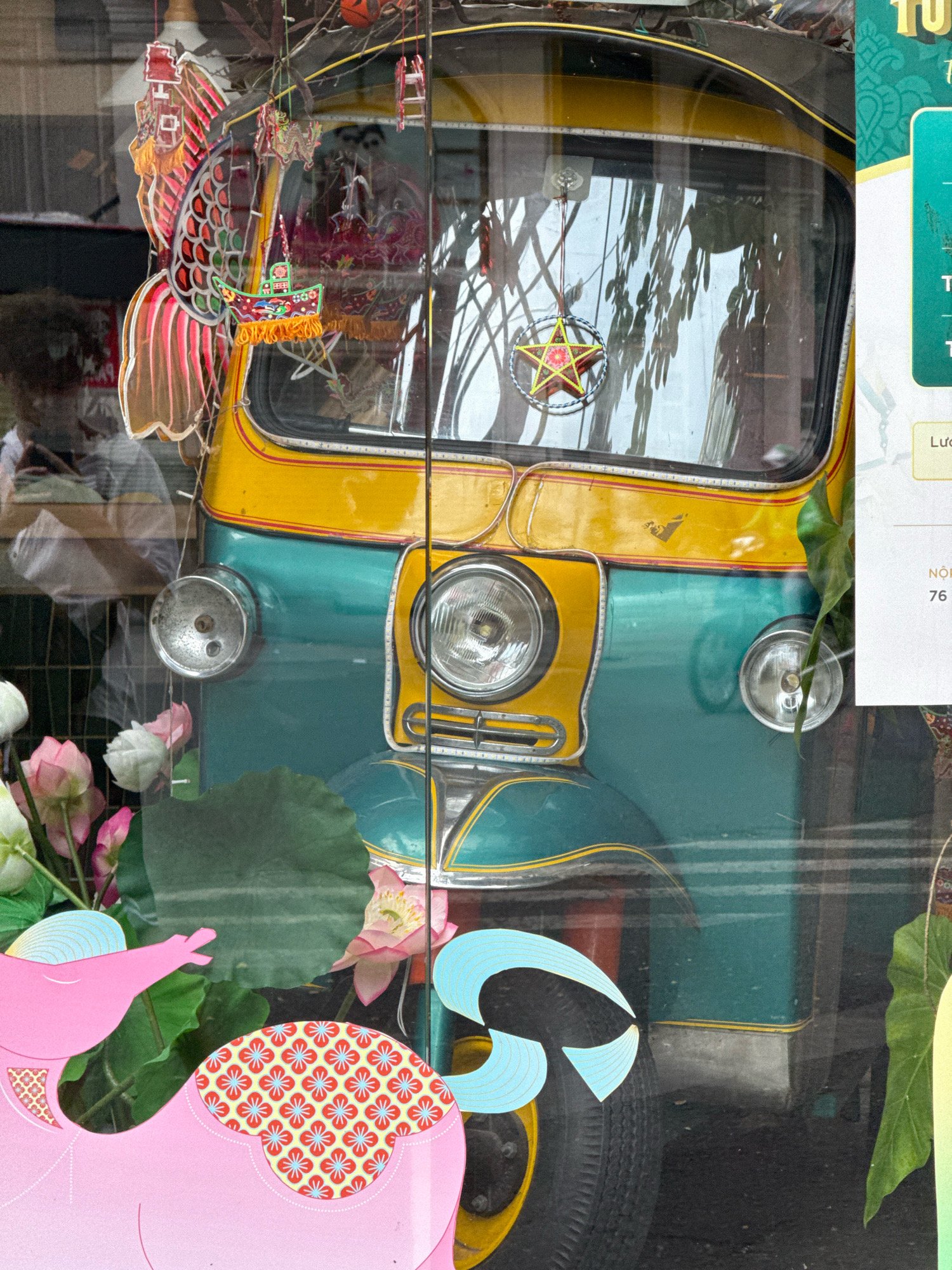 The image captures a close-up view of a colorful tuk-tuk, likely on a street in Nha Trang, Vietnam, seen through a glass window. The tuk-tuk features a bright yellow upper section with red and yellow stripes, and a teal lower body. It is adorned with various decorative elements hanging from its interior and exterior, including a large, ornate paper fish with red and pink scales, a star-shaped ornament, and what appears to be a small boat decoration with yellow fringe.
Reflected in the tuk-tuk's windshield are trees and possibly buildings, suggesting an outdoor urban setting. In the foreground, several artificial lotus flowers in shades of pink and white are visible, along with green lotus leaves. To the left and bottom of the image, large pink paper cutouts of elephants are present, one with a patterned saddle decorated with red and blue floral motifs.
A partially visible sign on the right side of the image, with green and yellow elements, appears to be a promotional or informational display. Text on the sign, though mostly obscured and in Vietnamese, includes the word "NGON" and a number sequence. The overall scene conveys a vibrant, festive, and possibly commercial atmosphere, with the tuk-tuk serving as a central visual element decorated for a special occasion or as a tourist attraction.