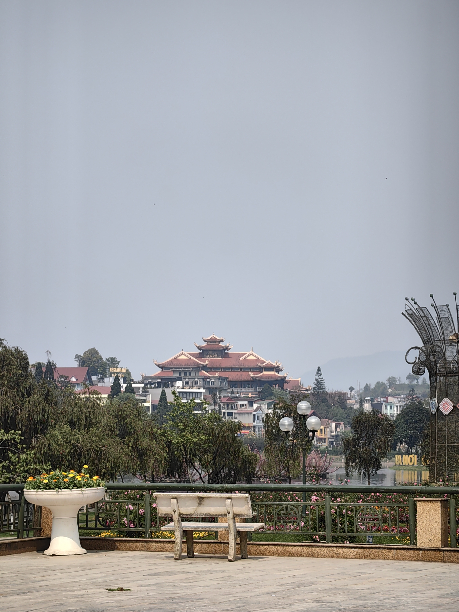 The image displays an outdoor scenic overlook in Sa Pa, Vietnam, presenting a view across Sa Pa Lake towards a hillside town. In the immediate foreground, a light-colored paved plaza features a white, pedestal-style concrete planter holding yellow and orange flowers, positioned on the left. To its right stands a single, light-toned concrete bench, facing away from the viewer towards a dark green metal railing adorned with decorative circular patterns. A light-colored rectangular trash receptacle is situated to the right, behind the bench, near the railing.
Beyond the railing, a landscaped garden area with pink and yellow flowering plants borders Sa Pa Lake. The lake's surface reflects the sky and parts of the distant bank, including a prominent golden sign spelling "HANOI." Numerous trees with dark green foliage are distributed along the lake's edge and up the hillsides, interspersed with multi-globed streetlights.
The background reveals a densely built town ascending a hillside. The most prominent structure is a large, multi-story building featuring multiple tiered roofs with red-orange tiles, indicative of traditional Vietnamese architectural style. Surrounding this central building are various other multi-story commercial and residential buildings with different roof colors. A sign identifying "RESTAURANT HA YEN" is visible on a building to the left of the dominant red-roofed structure. Distant mountains and hills are faintly discernible on the horizon through atmospheric haze. The sky overhead is overcast and light grey. No individuals are present, and no actions or interactions are depicted.