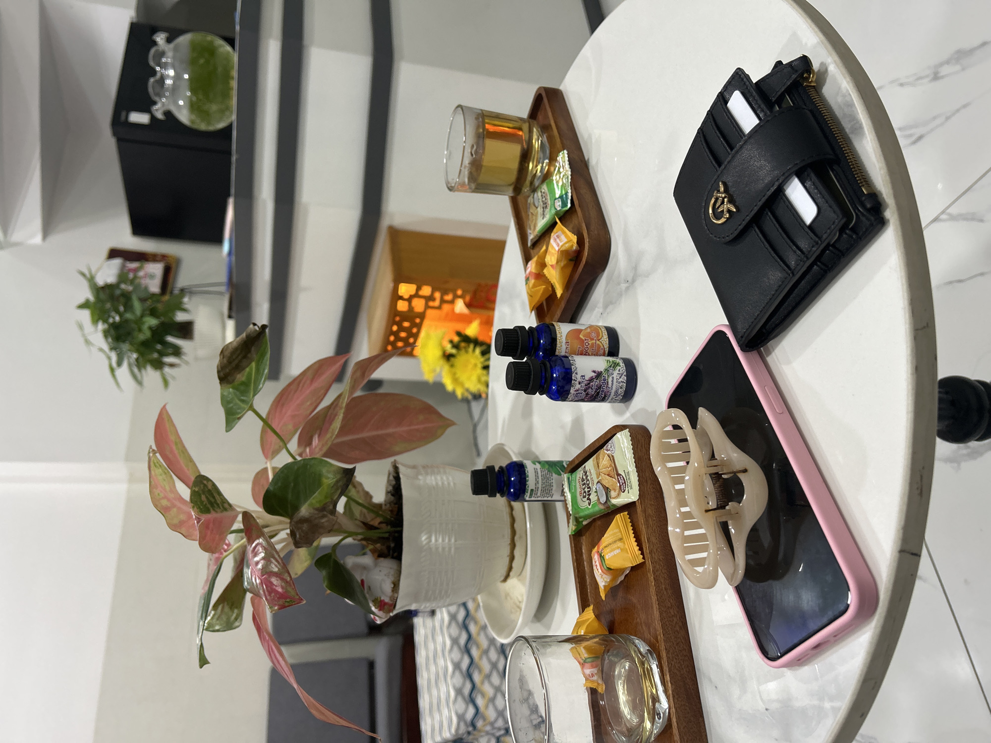 The media file displays a still life arrangement on a round, marble-patterned table, likely in a home or cafe setting in Nha Trang Ward, Vietnam. The scene is illuminated by indoor lighting, suggesting it could be daytime or evening.
Prominent objects include a black leather wallet with a gold logo and several cards peeking out, a pink-cased smartphone, a light-colored hair claw clip, a white potted plant with pink and green variegated leaves, and a small wooden tray holding a glass of amber liquid, a packet of tea or biscuits, and two individually wrapped snacks. Two small dark blue bottles, possibly essential oils or aromatherapy products, are placed near the tray. Another empty glass sits on the table closer to the viewer. In the background, a glimpse of a modern interior is visible with a black cabinet topped with a decorative glass container holding a green liquid, and a shelving unit with vertical gray stripes. Further in the background, a glimpse of yellow flowers and an orange-lit decorative screen can be seen through an opening.
There is no visible activity or event taking place; the arrangement appears staged or set for personal use. The overall mood is calm and serene, suggesting a moment of relaxation or preparation. Text is visible on the snack wrappers: "MANGO" is legible on one, and another appears to be "CRUNCH." On one of the blue bottles, the word "Lavender" can be partially read.