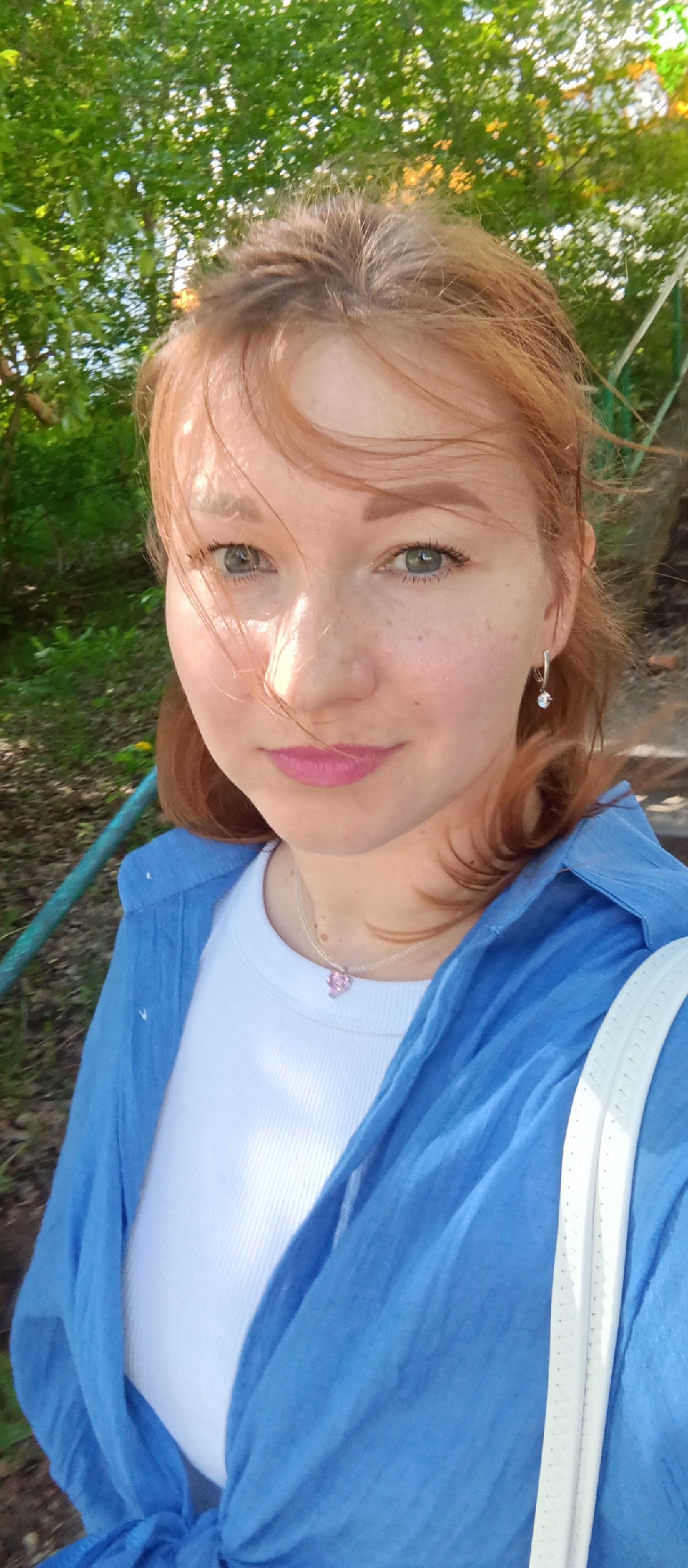 A close-up, eye-level shot shows a red-haired, fair-skinned woman with freckles, wearing a white t-shirt, a light blue button-down shirt, and a silver necklace with a pink charm. She is wearing silver hoop earrings with dangling crystals. The woman appears to be outdoors on a sunny day. The background consists of green foliage and parts of a blue metal railing, possibly indicating a park or garden setting. The specific location within the city of Unknown, Unknown cannot be determined from this image.