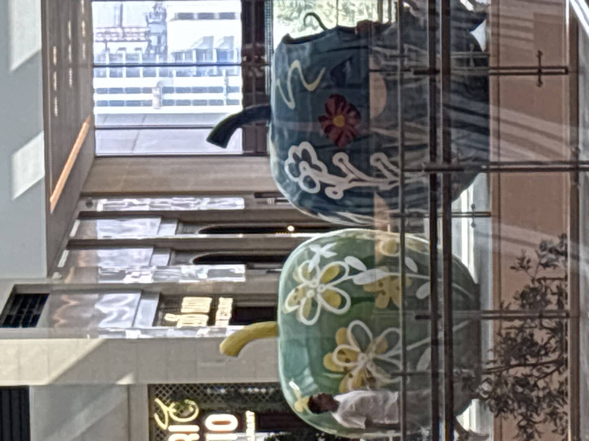 This image depicts two large, stylized apple sculptures inside a modern building, likely a mall or an atrium in Dubai, United Arab Emirates. The sculpture on the left is green with painted yellow and white flowers, while the one on the right is blue with white and red floral motifs. A man in a white t-shirt is visible standing near the green apple. The background features architectural elements of the building, including windows with a view of skyscrapers, and interior lighting. The sculptures are positioned behind a railing, suggesting they are part of an exhibition or decorative installation.
