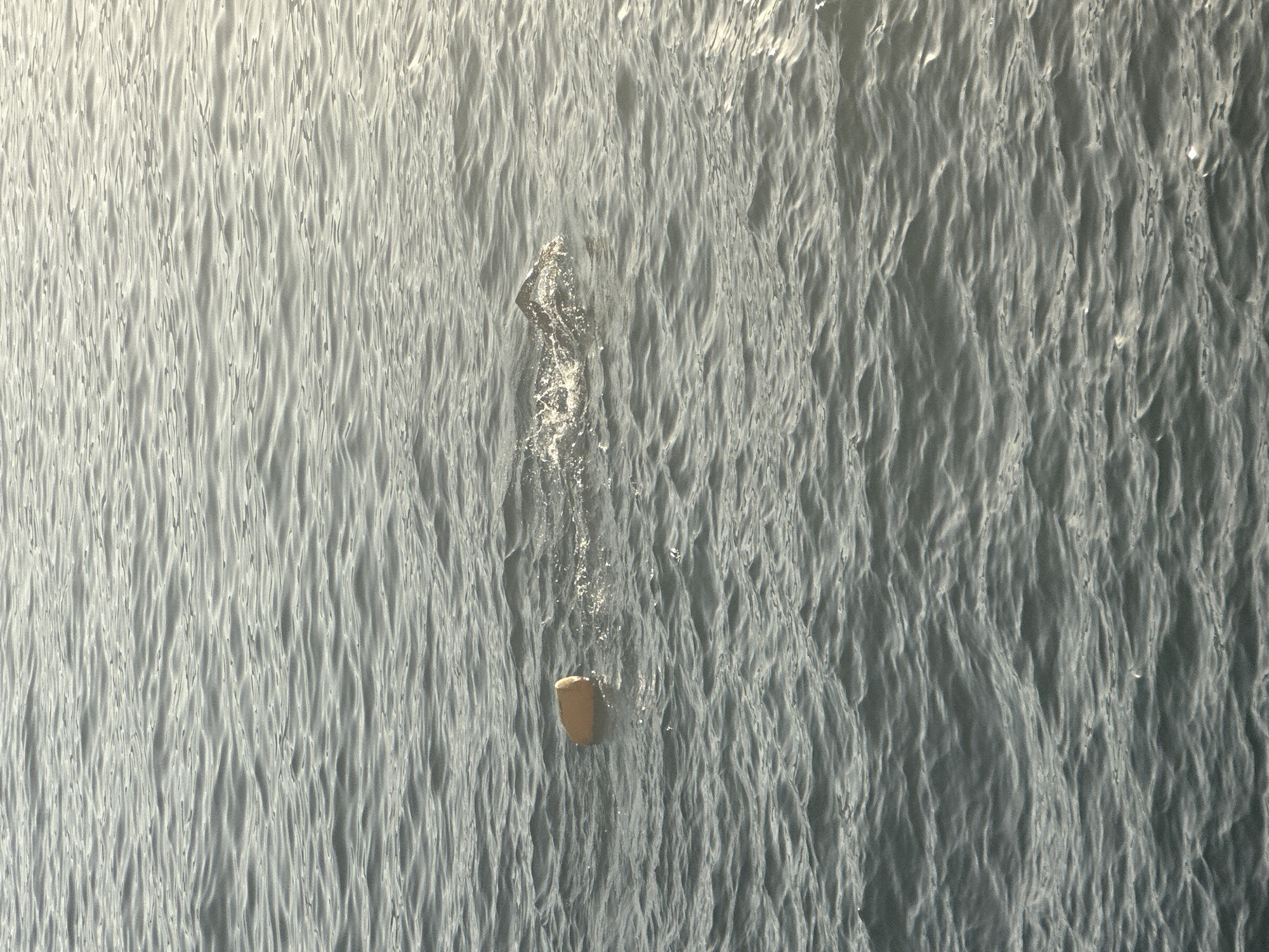 An orange buoy floats on the dark, choppy surface of the sea. Waves are visible across the entire image, with sunlight reflecting off the water's surface, particularly on the right side. The scene appears to be in the open water, likely off the coast of Budva, Montenegro, given the characteristic dark blue-grey sea and the presence of a navigational marker like a buoy. There are no people or buildings visible in the frame.