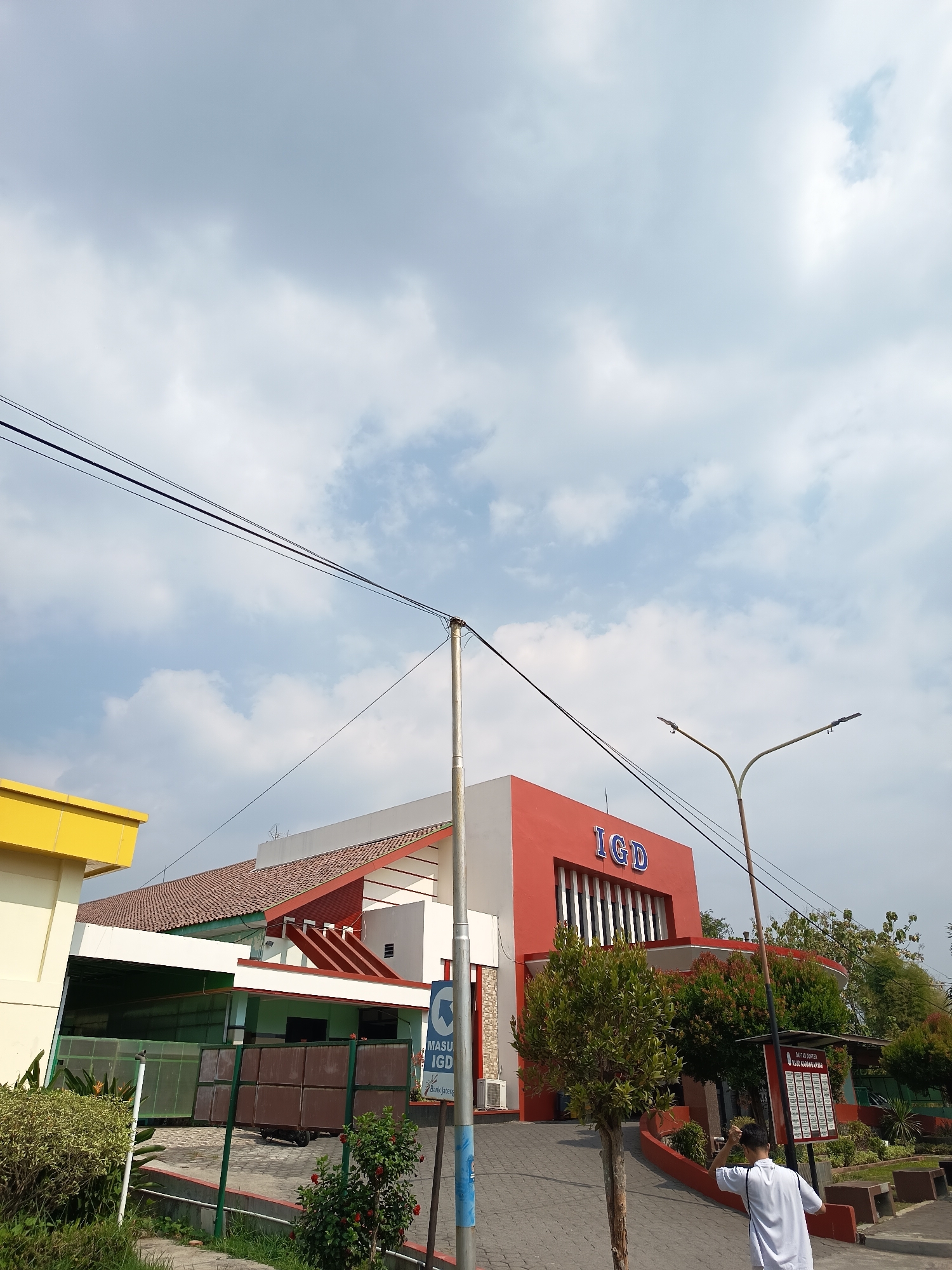 The image shows the exterior of a hospital building with a red facade and "IGD" written in blue letters. A yellow building is partially visible on the left. The sky is partly cloudy. In the foreground, a person in a white shirt is standing with their back to the camera, adjusting their hair. There are trees, bushes, and a street lamp in the scene. A sign with text in Indonesian indicates the location as "RSUD Karanganyar," which is a public hospital in Karanganyar, Indonesia. The scene appears to be an outdoor area of the hospital, possibly near the entrance.