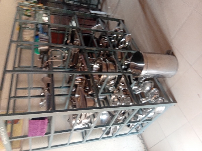 The scene is an interior view within Ejule, Nigeria, depicting a storage area for kitchen or catering equipment. A large, gray, multi-tiered metal shelving unit, approximately 2 meters high and 3 meters wide, dominates the image, extending from the left to the right side of the frame. This unit holds numerous items of reflective, clean stainless steel cookware and serving ware, including various sizes of pots, pans, bowls, platters, and ladles, systematically arranged on its multiple shelves. A folded purple towel is visible on the top left shelf. On the upper right shelves, several plastic containers and coiled hoses or cables are present. A cylindrical, stainless steel pedal-operated waste bin with a domed lid stands on the light-colored tiled floor in front of the lower right section of the shelving unit. The background wall is light-colored. No individuals are present, and no actions are observed.