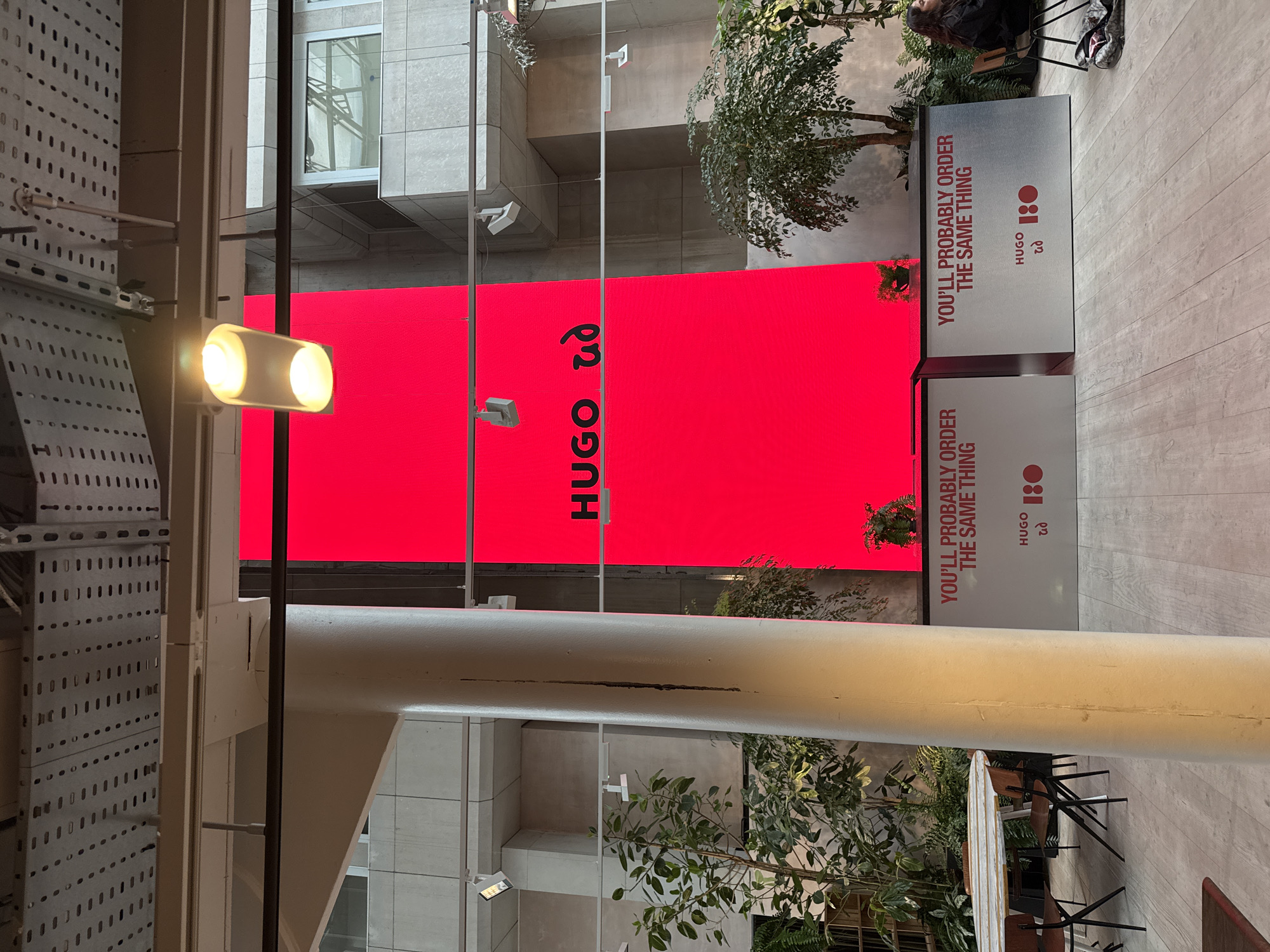 The image depicts a red HUGO advertisement in London, United Kingdom. The word "HUGO" is prominently displayed in large, bold lettering. Additionally, the phrase "YOU'LL PROBABLY ORDER THE SAME THING" appears on a sign near the ad. The sign also features the HUGO logo and "ad" written under it. The scene includes architectural details and greenery, suggesting an urban setting with modern design elements. One person appears sitting near the ads. The lighting is a combination of natural daylight and artificial light, creating a bright and somewhat abstract composition. The shot is taken from a low angle, emphasizing the height of the advertisements.