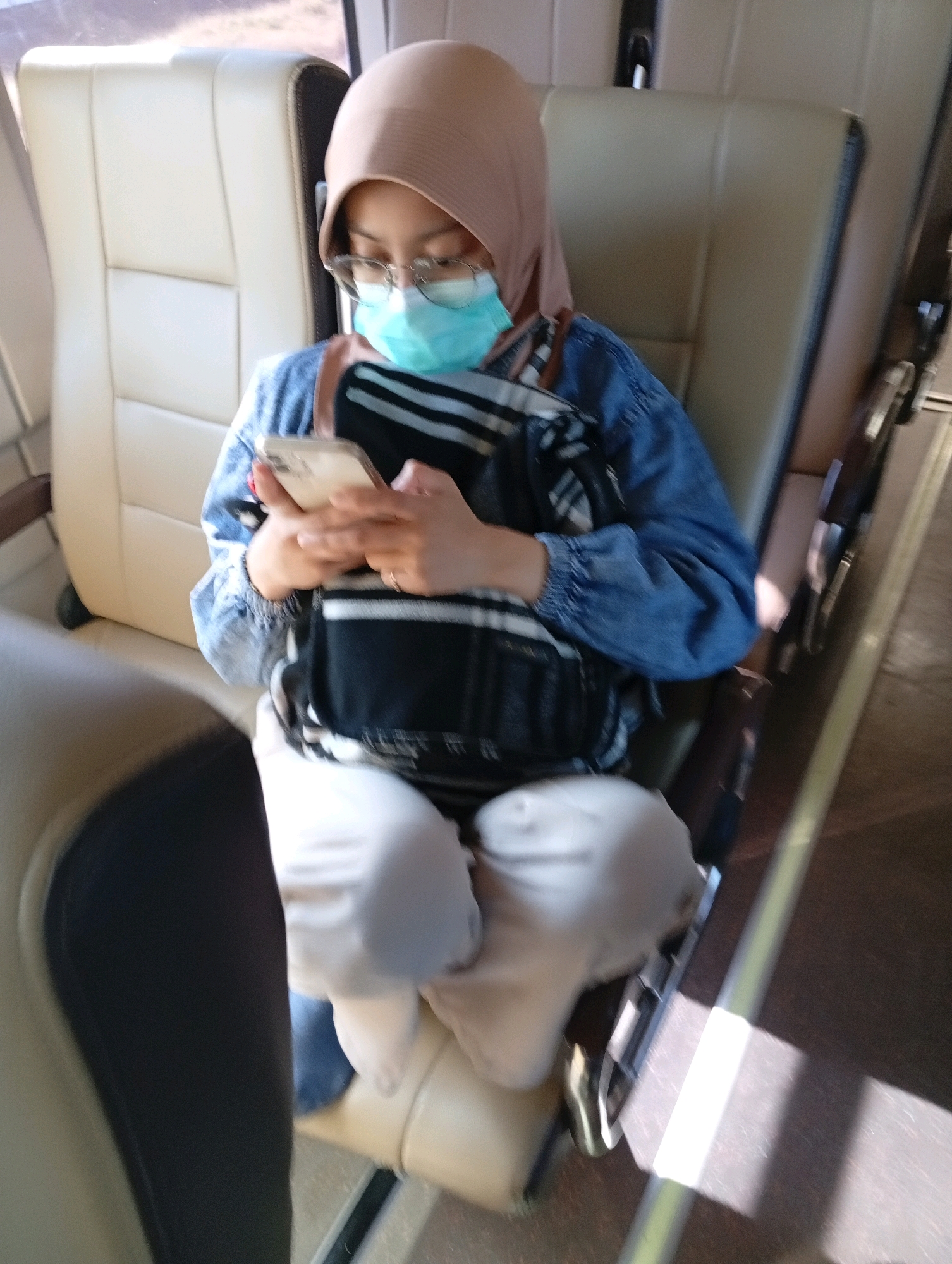 A woman wearing a light brown hijab, glasses, and a light blue face mask is seated on a tan colored vehicle seat, holding a smartphone and looking down at it. She is wearing a blue denim jacket over a black and white striped shirt, and light-colored pants. A black and white patterned bag is draped over her shoulders and across her chest. The seat is part of a row of similar seats, suggesting she is on public transportation, possibly a bus or train. The interior of the vehicle is visible, with another seat to her left and the aisle and seats to her right. Natural light is entering from the right side of the frame, creating bright highlights on the floor. The location is identified as Tebon, Indonesia.