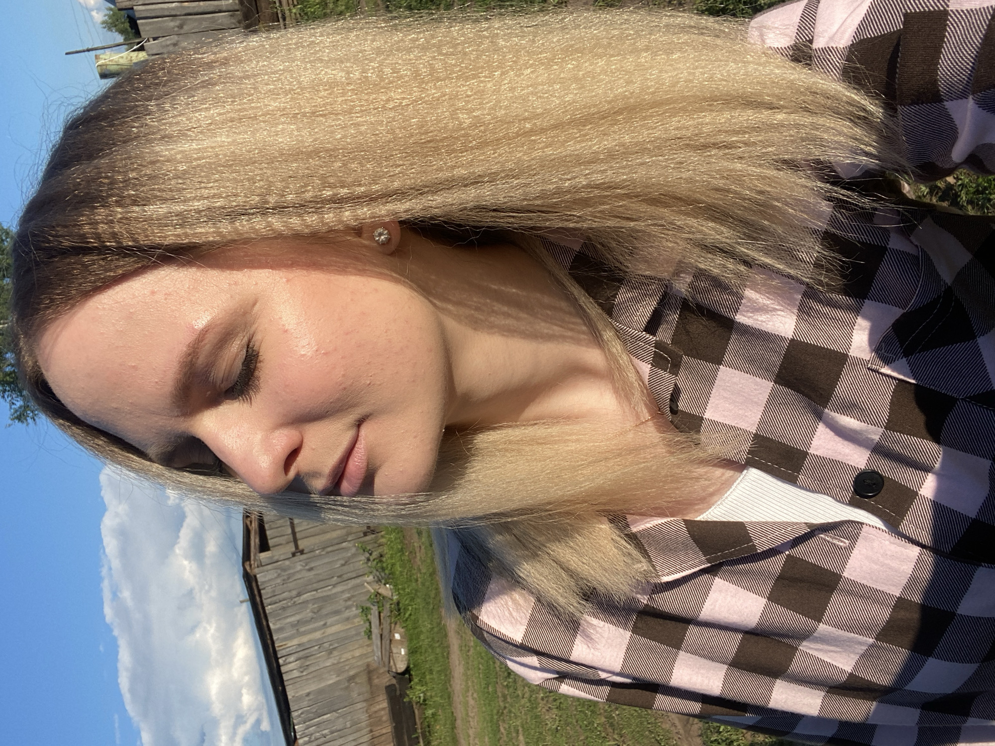 The image features a young woman outdoors in Kauchuk Stantsiya, Russia, under bright daylight conditions. Her head is tilted, and her eyes are closed or cast downwards, conveying a calm and contemplative expression. She has long, light blonde hair with visible darker roots, styled with a distinct crimped or textured appearance. The woman is wearing a pink and dark brown/black plaid button-up shirt, which is partially open to reveal a white ribbed top underneath. A small, sparkling stud earring is visible in her right earlobe.
Strong sunlight illuminates her hair and face, creating some shadows. The background indicates a rural or semi-rural setting, featuring a weathered wooden fence or building structure on the left, patches of green grass, and a clear blue sky dotted with white clouds. The time of day appears to be midday or afternoon due to the intensity of the light. No text is visible in the media.