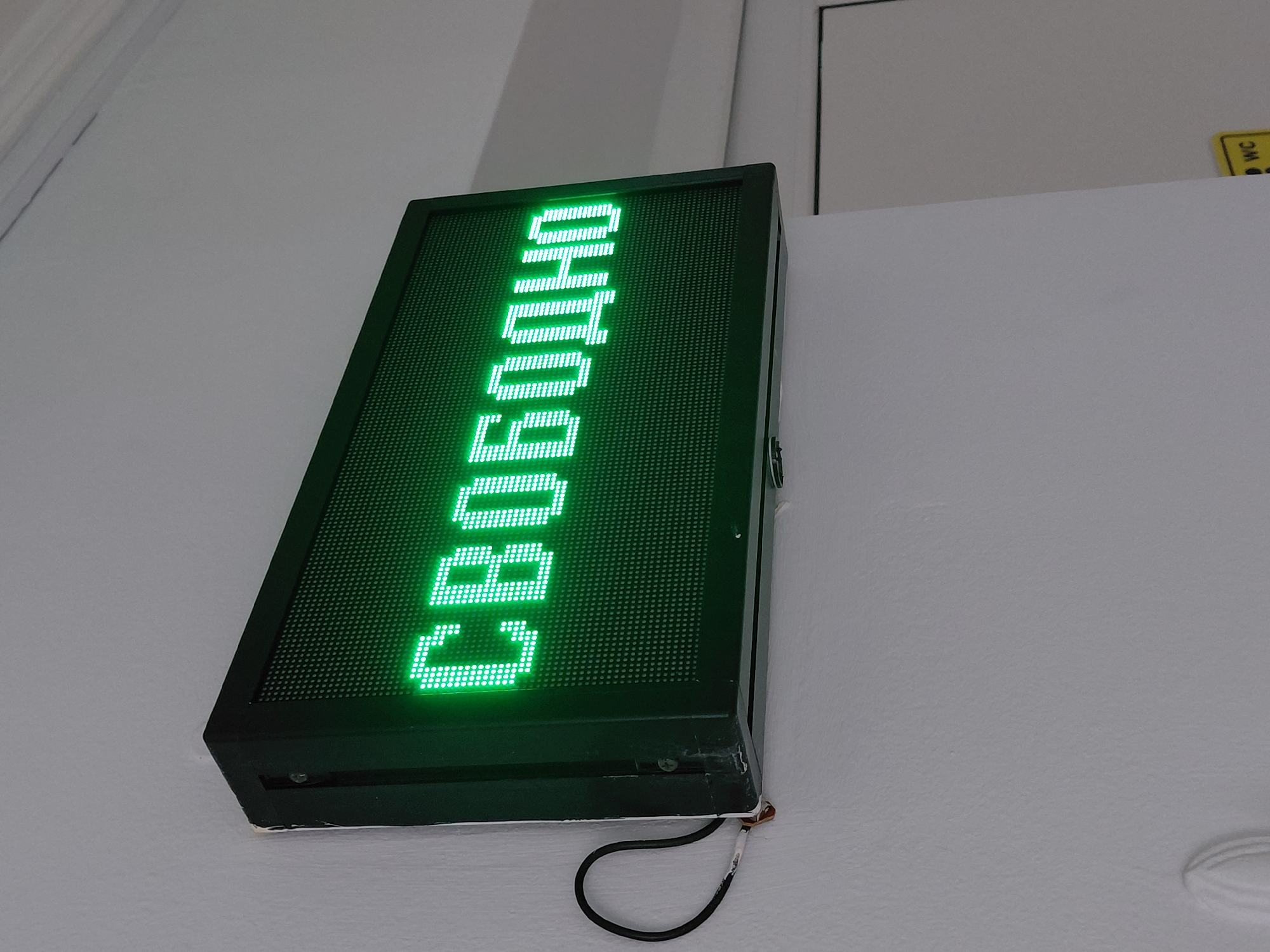 The image shows a green LED display mounted on a white wall. The display shows the Russian word "СВОБОДНО", which translates to "FREE" or "AVAILABLE" in English. The sign appears to be an indicator for a restroom or room availability. A black wire is visible at the bottom of the display. Part of a door and a small yellow sign with "WC" are seen in the background. The location of the image is Nadym, Russia.