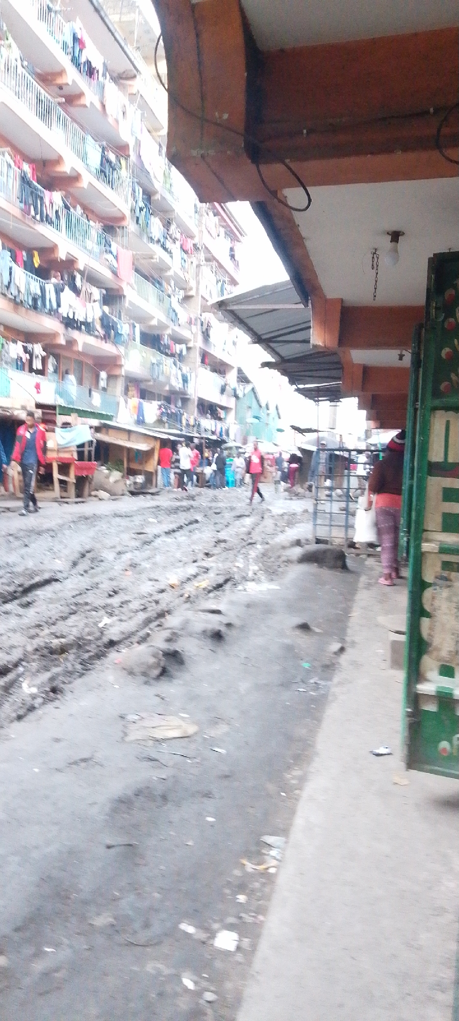 This media file captures a bustling street scene in what appears to be a residential and commercial area in Nairobi, Kenya. The focal point is a muddy, rutted road, indicative of poor drainage or recent rainfall.
On the left side of the image, a tall, multi-story apartment building stands prominently, its numerous balconies laden with drying laundry, suggesting a high-density residential environment. Below this building, several informal market stalls or small shops line the roadside.
The scene is populated by numerous individuals, mostly in the mid-ground. One person wearing a red jacket is visible on the left side of the muddy road, walking away from the camera. On the right, another person in a red top and patterned pants walks along a paved sidewalk. Other people are dispersed, going about their daily activities.
On the right side of the frame, a structure with a sturdy wooden overhang and beams is visible, beneath which a corrugated metal awning provides cover. A single light bulb hangs from the ceiling of this structure. A partial view of a green, possibly metal, structure, perhaps a shop door or shutter, is seen on the far right, displaying some indistinct and illegible white text.
The lighting suggests it is daytime, possibly under an overcast sky given the lack of strong shadows. The overall impression is one of everyday life in a densely populated urban setting, characterized by active street commerce and residential living in close proximity.