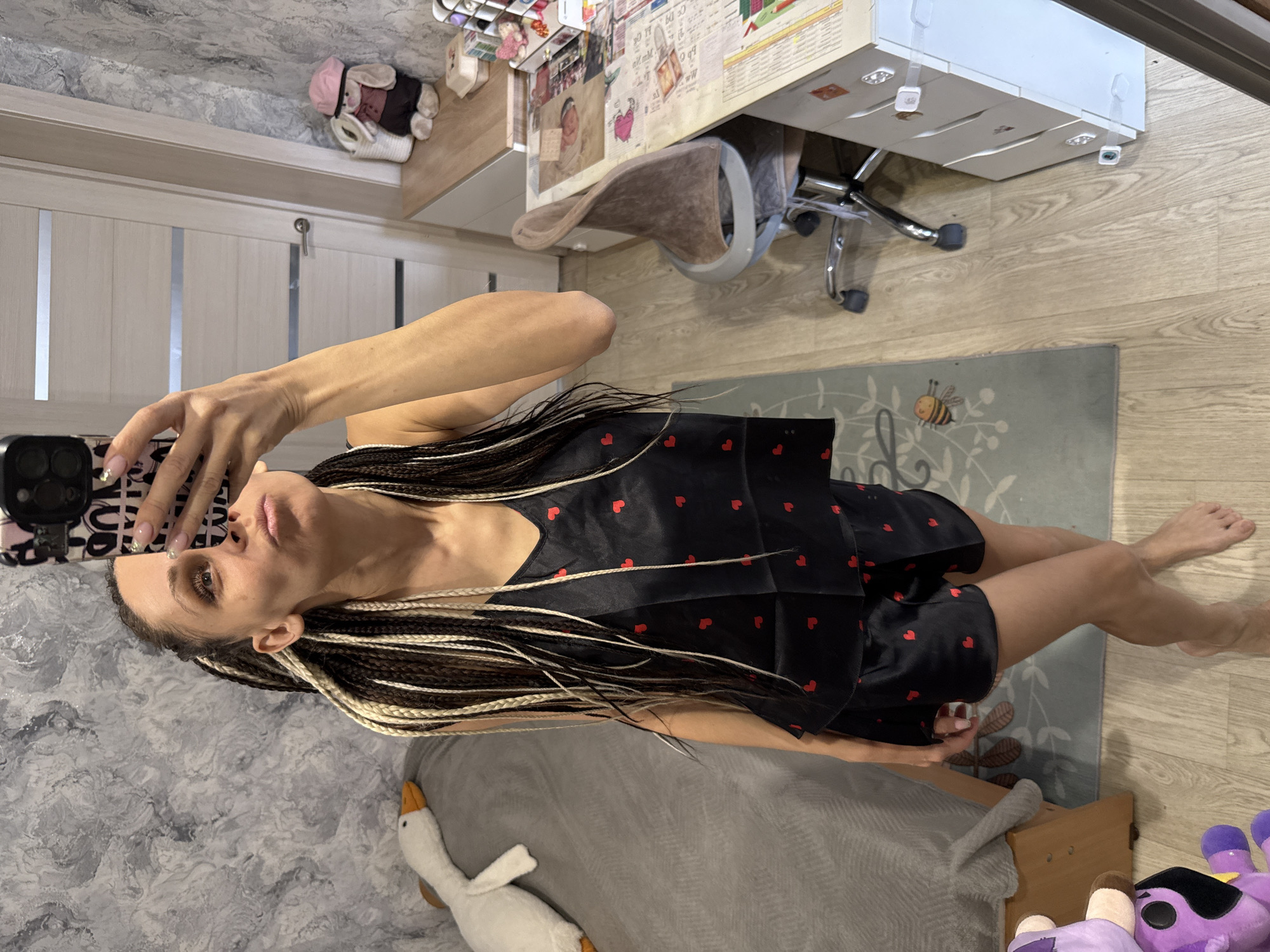 A young woman, appearing to be in her 20s or 30s, takes a selfie in what appears to be a bedroom or personal living space in Rusanovo, Russia. She holds a smartphone with a black camera module and a white and pink patterned case (possibly reading "LOVE") up towards herself. Her expression is neutral. She has long, multi-toned braided hair (dark brown/black and blonde) and is wearing black pajamas with small red heart patterns. Her bare feet are visible on the floor.
The room features a light-colored wooden floor and a grey rug with a cartoon bee and leaf design. To the left, a bed with grey bedding holds a white stuffed goose toy. A purple stuffed toy is visible on the floor near the bed. In the background, a light-colored wooden desk with a white drawer unit underneath is cluttered with papers, notes, and possibly small personal items. A brown fabric desk chair with a metal frame is tucked under the desk. The walls are partly visible; one wall has a grey, marble-like texture, while another is a plain light color. A light-colored wooden door with vertical panels is also visible. Above the door, a small shelf holds neatly folded white towels and a plush toy wearing a pink hat. The overall lighting suggests it is daytime or evening indoors.