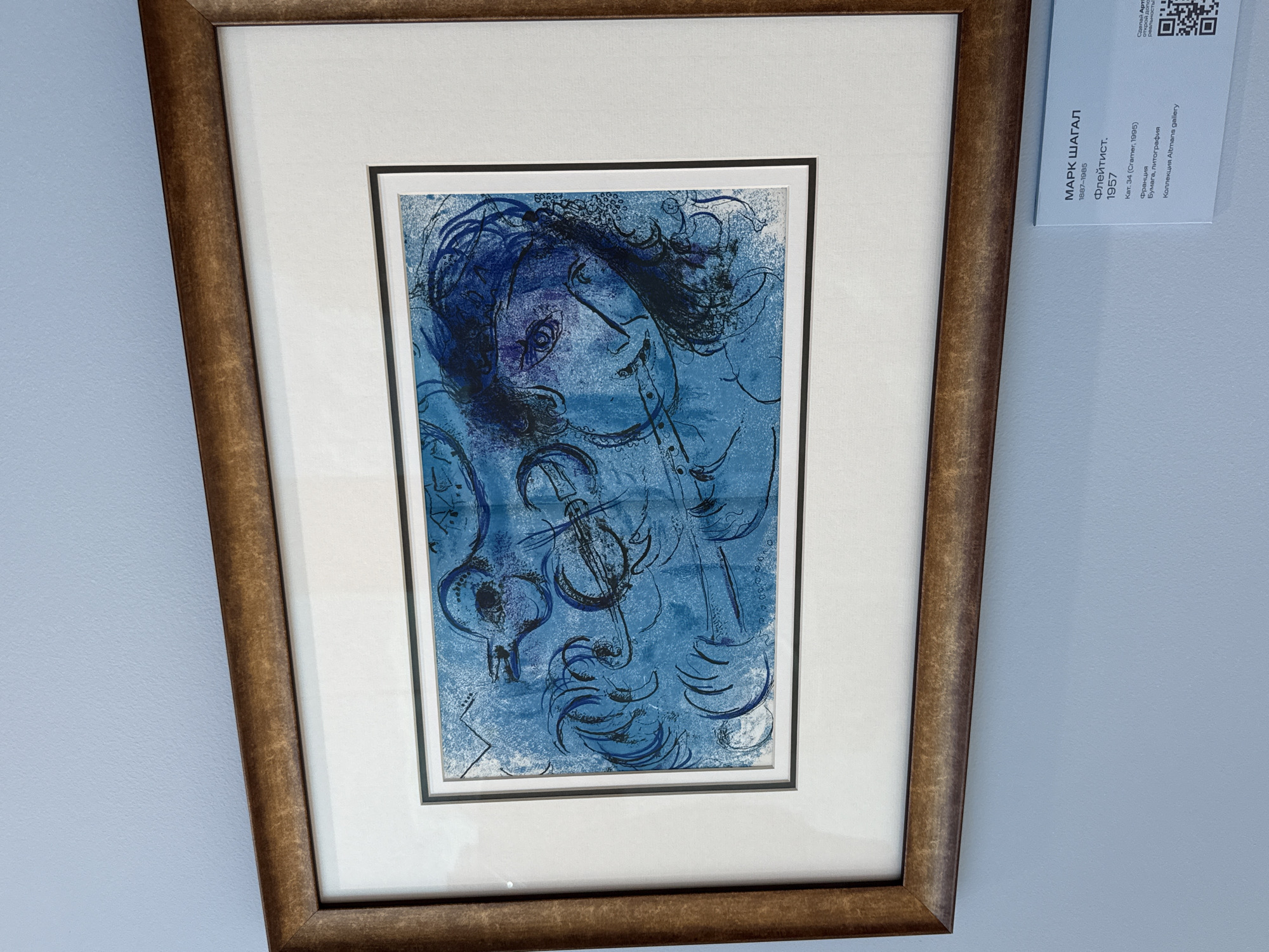 The image shows a framed artwork hanging on a light blue wall. The artwork is a blue lithograph featuring a stylized depiction of a person playing a flute, with musical instruments and abstract shapes incorporated into the composition. The frame is made of wood with a distressed, golden-brown finish. Below the artwork, a white placard with black text provides details about the piece, including the artist's name "МАРК ШАГАЛ" (Marc Chagall), dates of life "1887-1985", title "Флейтист." (The Flutist), year "1957", catalog number "Кат. 34 (Cramer, 1995)", country of origin "Франция" (France), medium "Бумага, литография" (Paper, lithography), and collection "Коллекция Altmans gallery". A QR code is also visible on the right side of the placard. The lighting suggests an indoor environment, possibly a gallery or museum. The overall scene is clean and well-lit, with the artwork serving as the central focus.