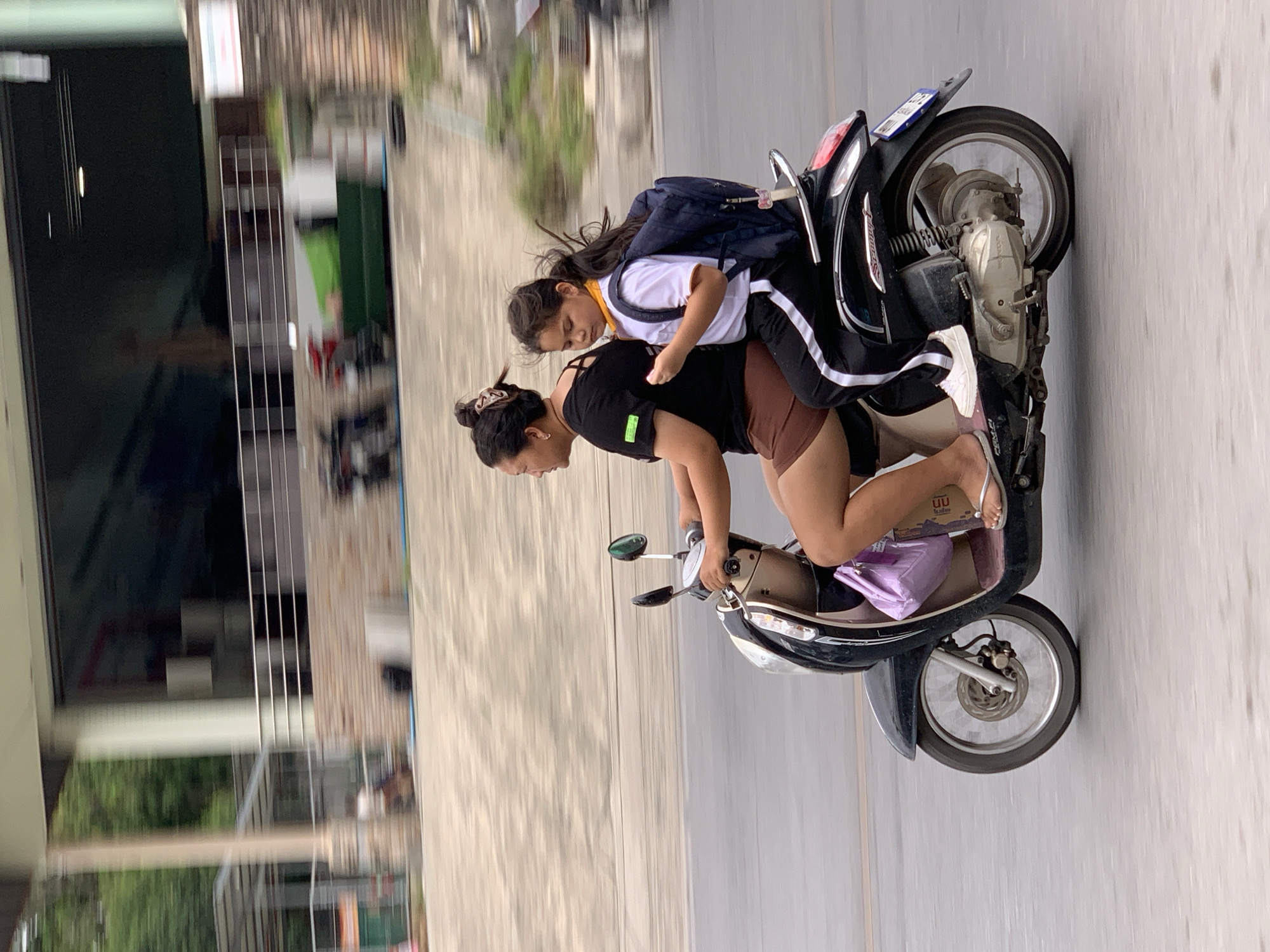 A woman and a young girl ride on a black scooter on a street in Ko Samui District, Thailand. The woman, who is driving, wears a black t-shirt and brown shorts. The girl, seated behind her, wears a white t-shirt, black pants with a white stripe, and a blue backpack. She has a yellow bandana around her neck. The scooter is a dark color with silver accents. In the background, there is a blurred building with a dark glass facade and a beige-colored wall. Trees and other structures are also visible, all blurred due to motion. The road surface is gray asphalt.