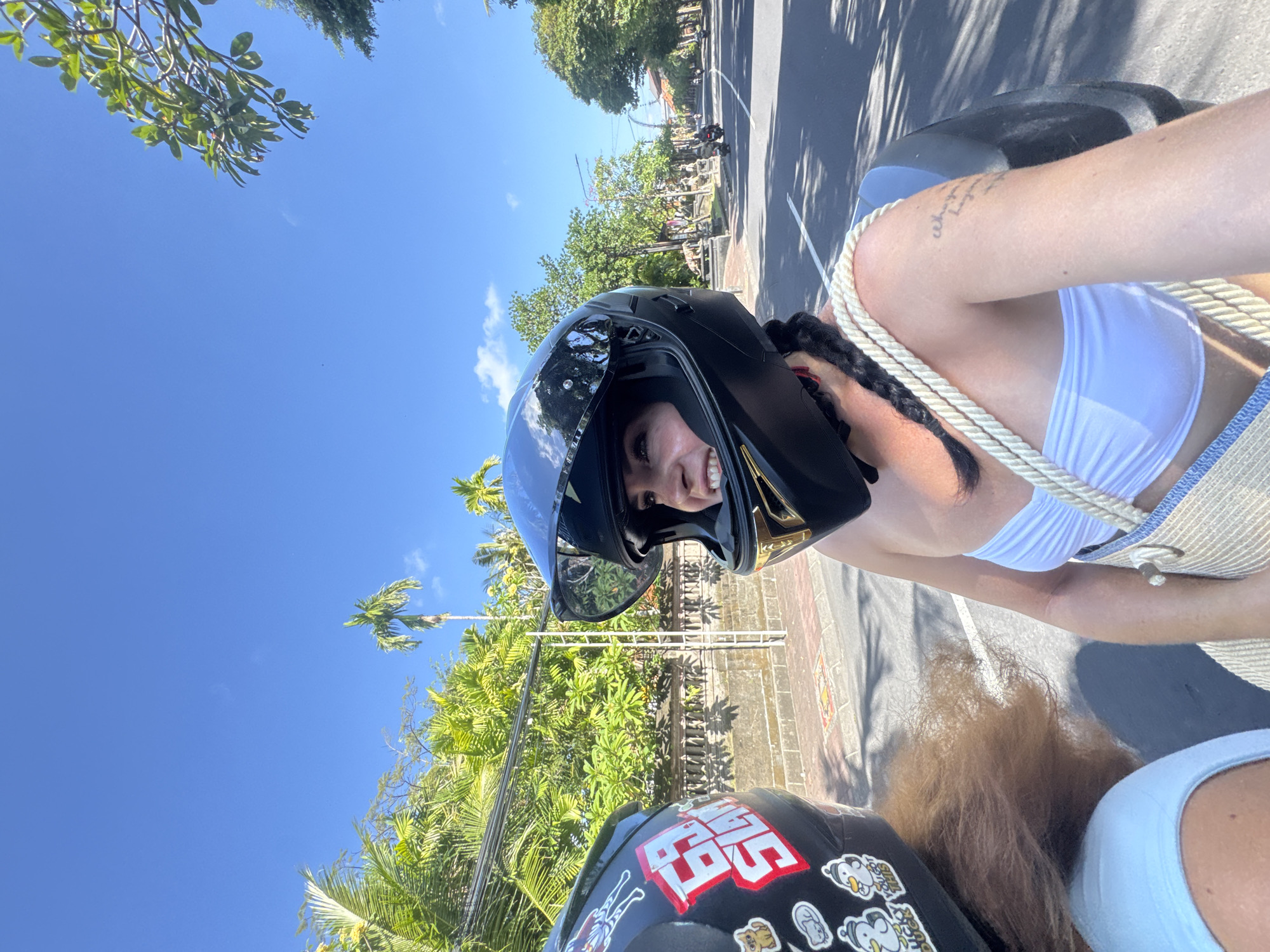Two people, wearing helmets and riding on a scooter, are traveling down a street lined with palm trees and lush greenery in North Kuta, Indonesia. The person in the foreground is a woman with a black helmet, white strapless top, and a woven straw bag slung over her shoulder. Her hair is braided and hangs over her shoulder. She is looking to her right, smiling. The other person, whose helmet is partially visible on the left, appears to have brown hair. The street is paved with sidewalks and a wall with ornate carvings runs along the left side of the road. Other scooters and motorcycles can be seen in the distance. The sky is clear and blue with a few wispy clouds.