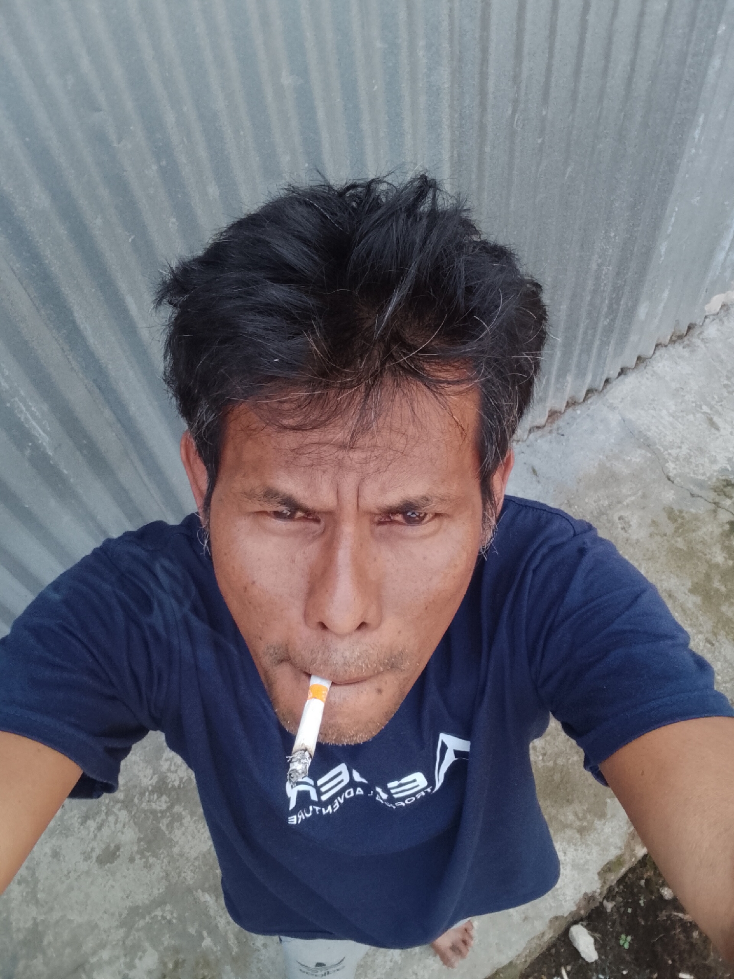 In an outdoor setting, a man is taking a selfie. He is wearing a dark blue t-shirt with a white logo on the chest, which appears to say "ADVENTURE" in reverse. A lit cigarette is held between his lips. His dark hair is disheveled, and his face is turned towards the camera, with a furrowed brow. Behind him, a corrugated metal wall forms the backdrop, and the ground is a mix of concrete and dirt. The overall scene suggests a casual, possibly rural or semi-urban environment in Padang Dalom, Indonesia.