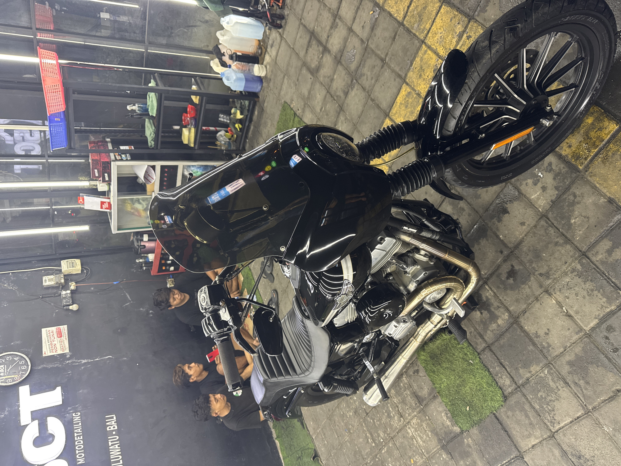 Here is a detailed description of the scene:
The image shows a black motorcycle parked indoors. The location is a "MOTODETAILING" shop, specifically "OCT MOTODETAILING ULUWATU - BALI" indicated by a sign on the wall. Uluwatu is located in Ungasan, Indonesia.
Several men are present in the shot; they seem to be examining or detailing the motorcycle. The motorcycle has a sleek design, and its front wheel is turned slightly to the right. The floor is tiled, and there are patches of artificial grass surrounding the motorcycle. Shelves filled with cleaning supplies are visible in the background. The shop appears well-lit and organized.